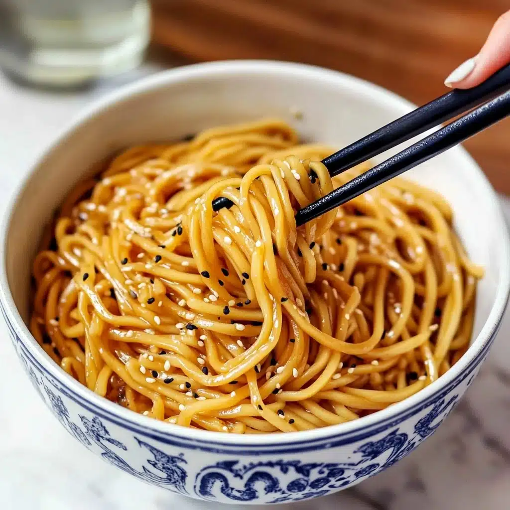 Hibachi Noodles, Hibachi Noodles recipe, easy hibachi noodles, homemade hibachi noodles, restaurant-style hibachi noodles A bowl with blue and white patterns holds a serving of yellow noodles coated in a shiny sauce, with black and white sesame seeds sprinkled on top. Two wooden chopsticks with black grips grasp a small bundle of noodles above the bowl. The bowl is placed on a brown wooden surface with a white marbled background partly visible.