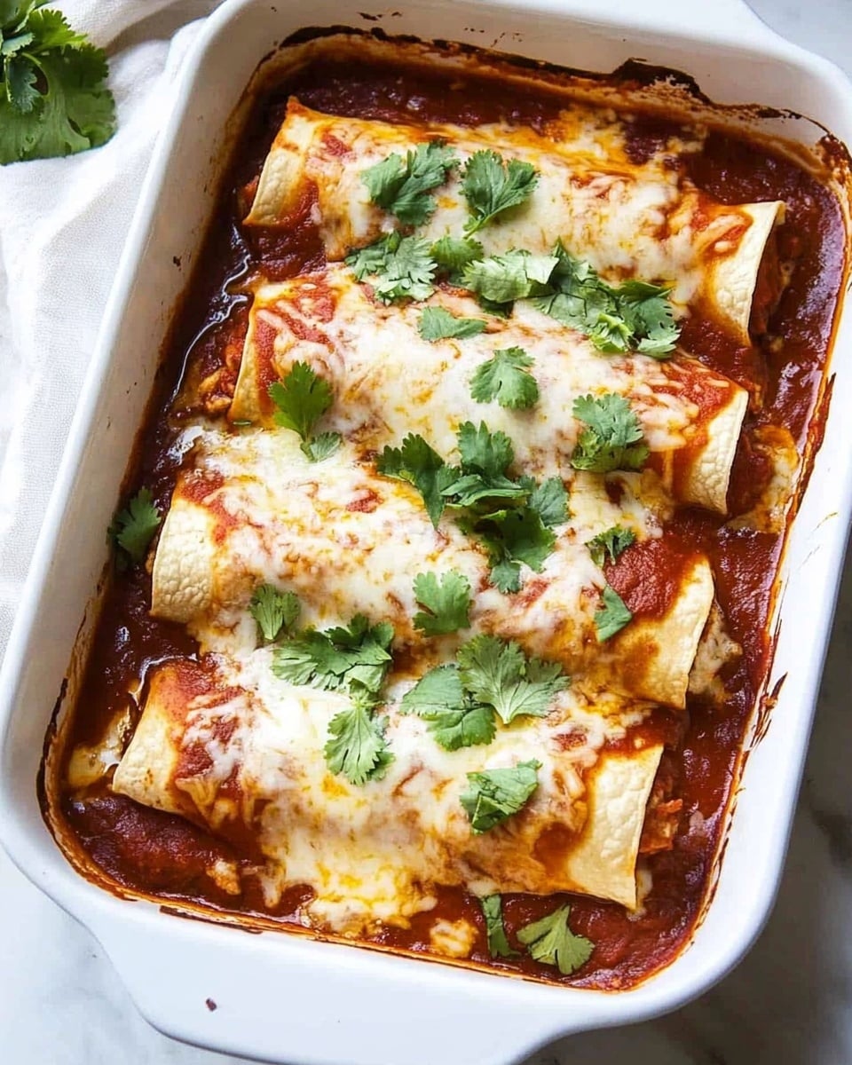 Beef Enchiladas, Beef Enchiladas Recipe, Easy Mexican Enchiladas, Homemade Beef Enchiladas, Comfort Food Enchiladas A white rectangular baking dish filled with four rolled tortillas placed side by side, covered in a thick dark red sauce. The tortillas are topped with a layer of melted pale yellow cheese that looks soft and stringy. Fresh green chopped cilantro is sprinkled evenly over the cheese, adding a fresh contrast in color. The dish is set on a white marbled surface with a white cloth partially visible on the left side.