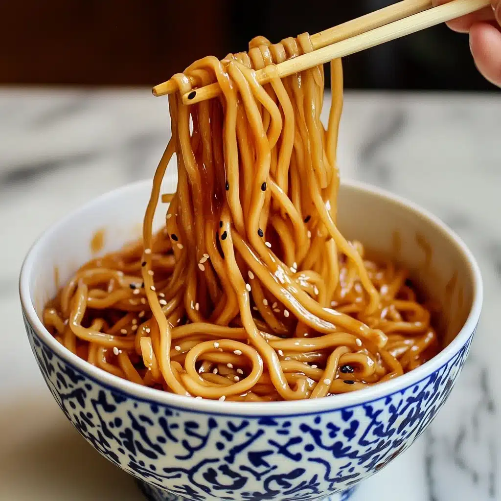 Hibachi Noodles, Hibachi Noodles recipe, easy hibachi noodles, homemade hibachi noodles, restaurant-style hibachi noodles A white bowl with blue patterns holds a large serving of brown noodles mixed with black and white sesame seeds. The noodles are thick and shiny, piled high inside the bowl with some strands lifted by a pair of wooden chopsticks held by a woman's hand. In the background, there are blurred bottles and a white cloth on a white marbled surface.