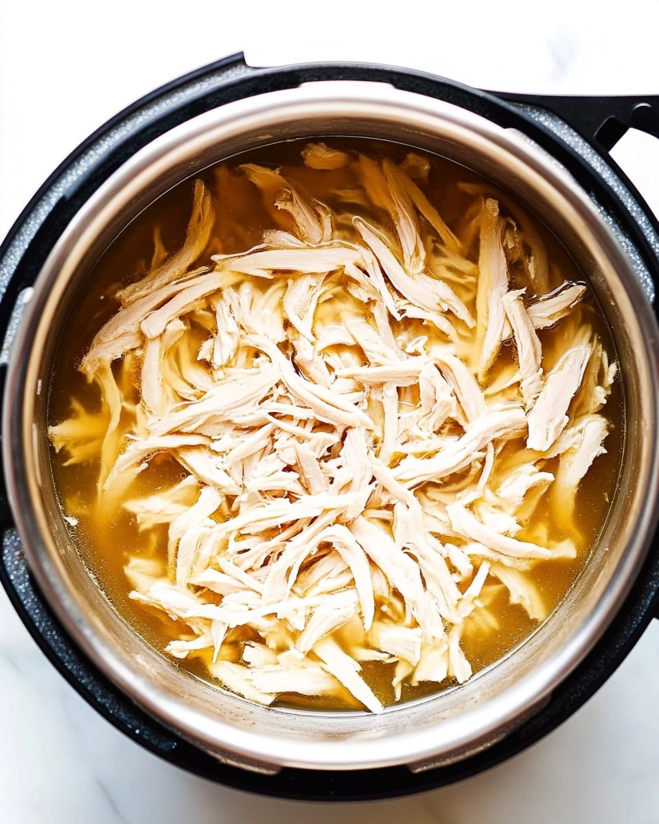 The image shows a close-up of shredded pale beige chicken pieces resting in a shiny silver pot filled with light brown broth. The tender chicken strands are scattered unevenly throughout the liquid, creating a mix of soft textures and moist layers within the pot. The pot itself has a black outer rim and handle, set on a white marbled surface. The lighting highlights the moistness and slight sheen of the chicken and broth. Photo taken with an iphone --ar 4:5 --v 6.1