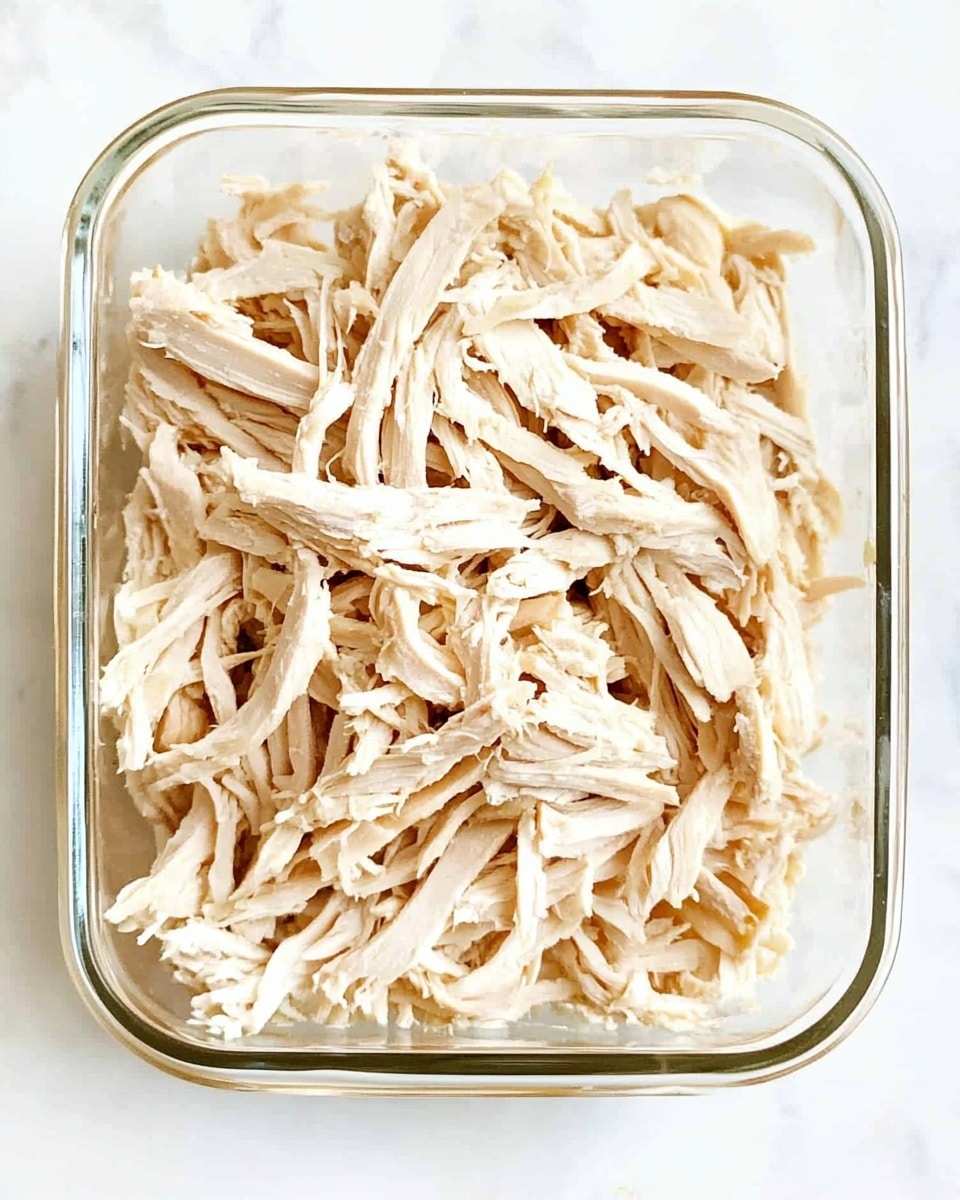A rectangular glass container filled fully with shredded light tan chicken pieces, showing texture from the torn strands stacked evenly. The chicken looks moist and soft, with light shadows adding depth inside the clear container. The container sits on a white marbled surface, with no other objects visible. Photo taken with an iphone --ar 4:5 --v 6.1