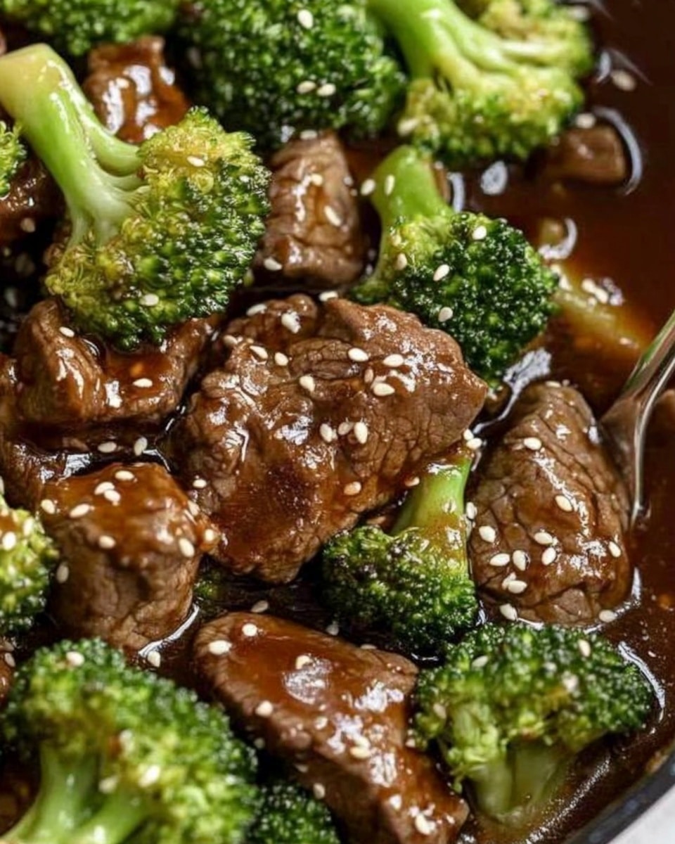 Chinese Beef and Broccoli Stir Fry, Chinese beef and broccoli, beef stir fry recipe, Chinese takeout at home, quick beef broccoli dish The dish shows a white plate filled with a base layer of fluffy white rice. On top, there is a mix of bright green broccoli florets and cooked slices of brown beef coated in a glossy, thick brown sauce. The broccoli looks fresh and slightly steamed, while the beef pieces have a tender, slightly shiny texture. White sesame seeds are sprinkled over the beef and broccoli, adding a light touch of texture. A pair of wooden chopsticks rests on the edge of the plate, which is placed on a white marbled surface.