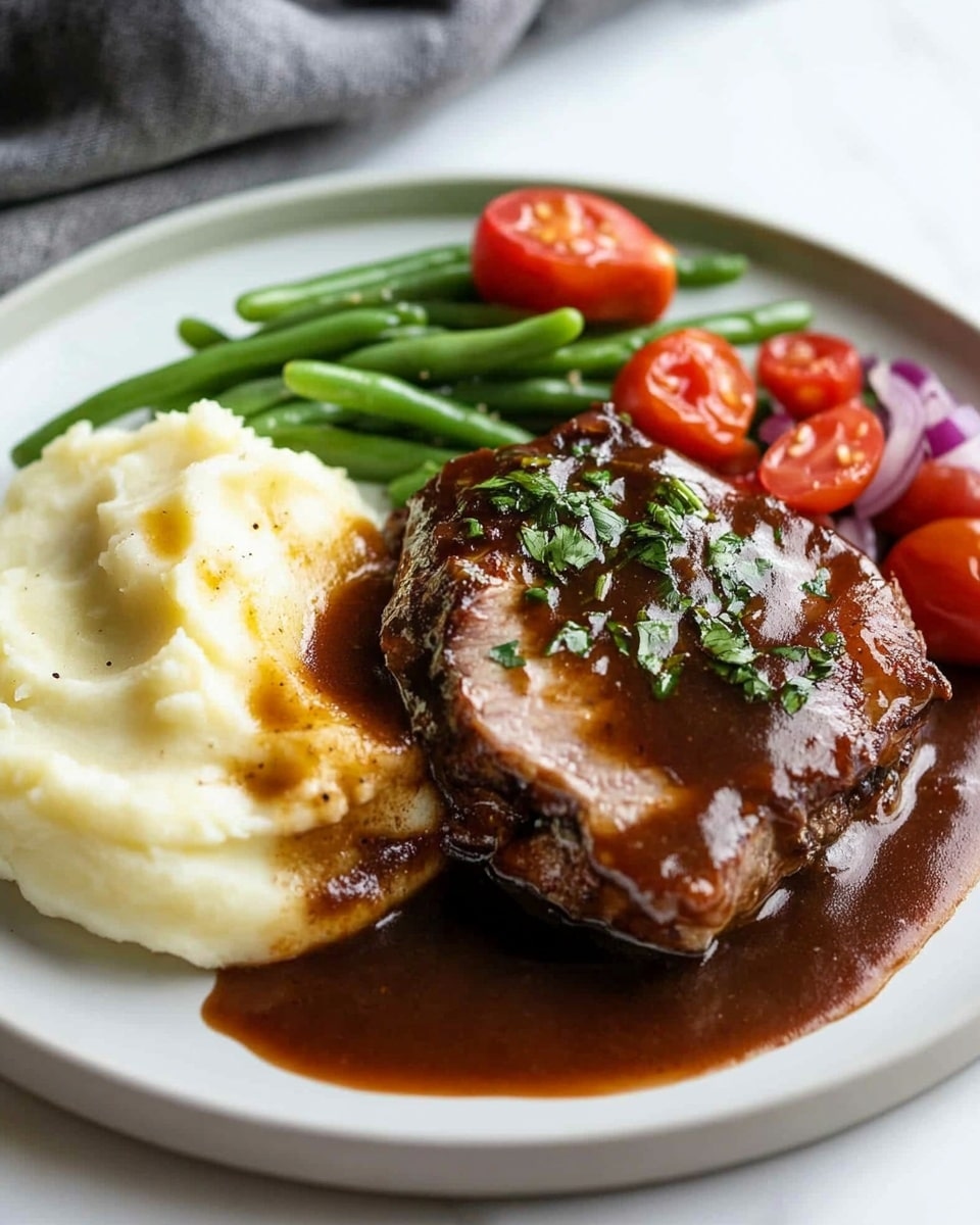 Oven Baked BBQ Chicken, Quick Homemade Barbecue Sauce, Easy BBQ Chicken Dinner, Juicy Oven Baked Chicken, Simple BBQ Chicken Recipes A close-up view of a thick piece of meat with a shiny brown sauce on top, sprinkled lightly with small green herb pieces. The meat sits on a base of creamy, light yellow mashed potatoes that look soft and smooth. To the side, bright red cherry tomatoes and green beans add color in the background, all on a white plate with a white marbled surface underneath. A fork holds a small slice of the meat, showing its juicy inside.