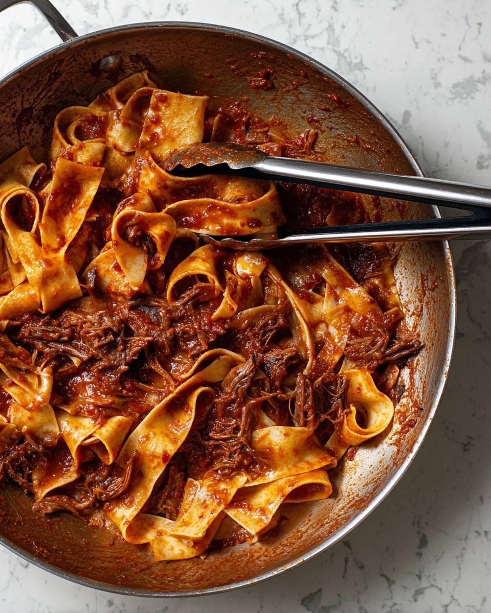 Inside a large pan with a metallic surface, there are wide, flat pasta noodles coated with a rich, deep red tomato sauce. The sauce has a thick texture and contains pieces of shredded dark brown meat. The noodles are glossy and curled, sitting unevenly across the pan's bottom. Two metal tongs with black handles rest across the pan, partially covering the pasta. The scene is set on a white marbled surface. Photo taken with an iphone --ar 4:5 --v 7