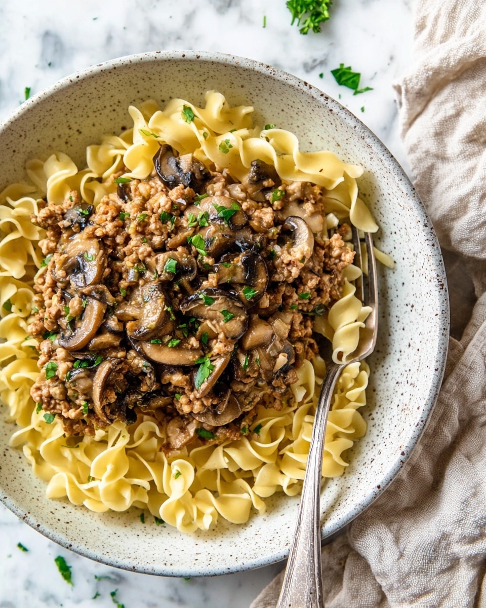 A white speckled bowl holds a dish with two visible layers: the bottom layer is a bed of light yellow egg noodles with a slightly curled and soft texture, arranged loosely to fill the bowl's base. The top layer is a creamy, light brown sauce mixed with ground meat, sautéed translucent onion pieces, and dark brown cooked mushroom slices, all evenly spread over the noodles. Small green parsley leaves are sprinkled on top for color contrast. A fork rests inside the bowl with some noodles and sauce on it. The bowl is set on a white marbled surface with a beige cloth partially visible in the background. photo taken with an iphone --ar 4:5 --v 7