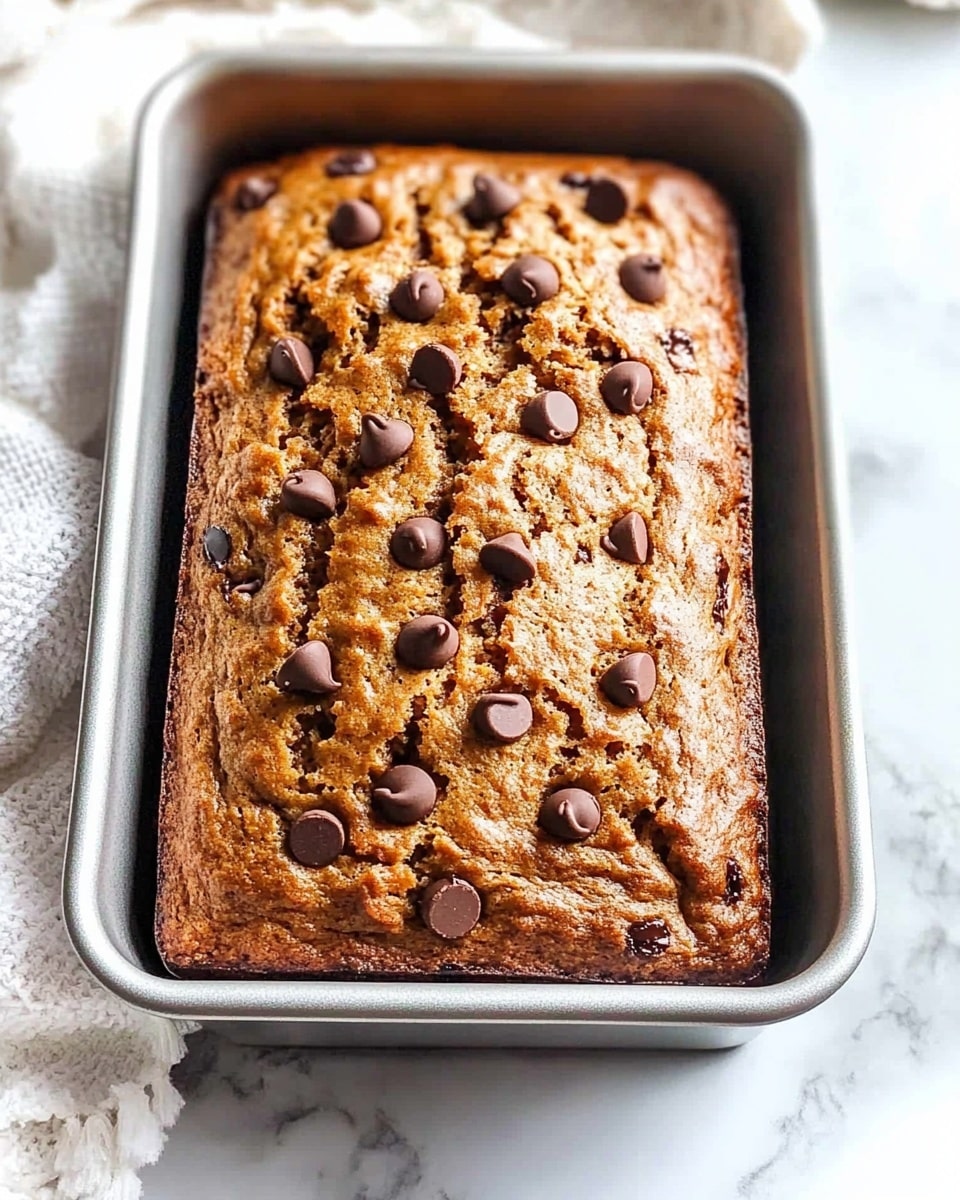 A golden-brown baked loaf with a slightly cracked top, studded generously with dark chocolate chips spread across the surface, sits inside a metal rectangular baking pan lined on both sides with parchment paper. The texture looks soft and slightly crumbly, with the chocolate chips melted just a bit but keeping their shape. The pan rests on a white marbled surface with a part of a white cloth visible on the side. photo taken with an iphone --ar 4:5 --v 6.1