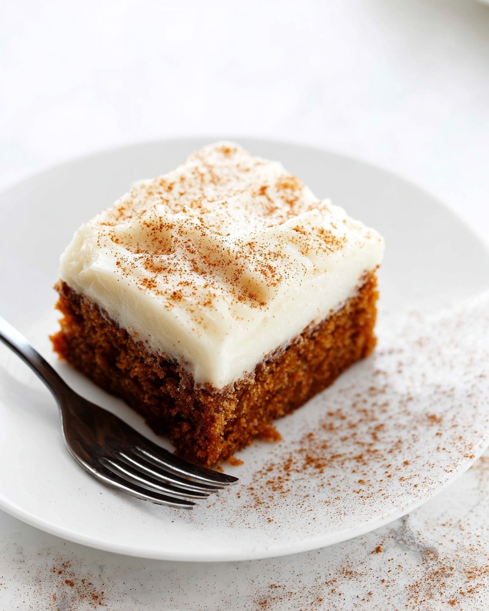 A square piece of carrot cake sits on a white plate with a silver fork resting beside it. The cake has two visible layers: a thick, moist, textured bottom layer with a warm brown color, and a smooth, creamy white frosting layer on top, dusted lightly with fine brown spice powder. The plate rests on a white marbled surface, adding softness and brightness to the image. Photo taken with an iphone --ar 4:5 --v 7