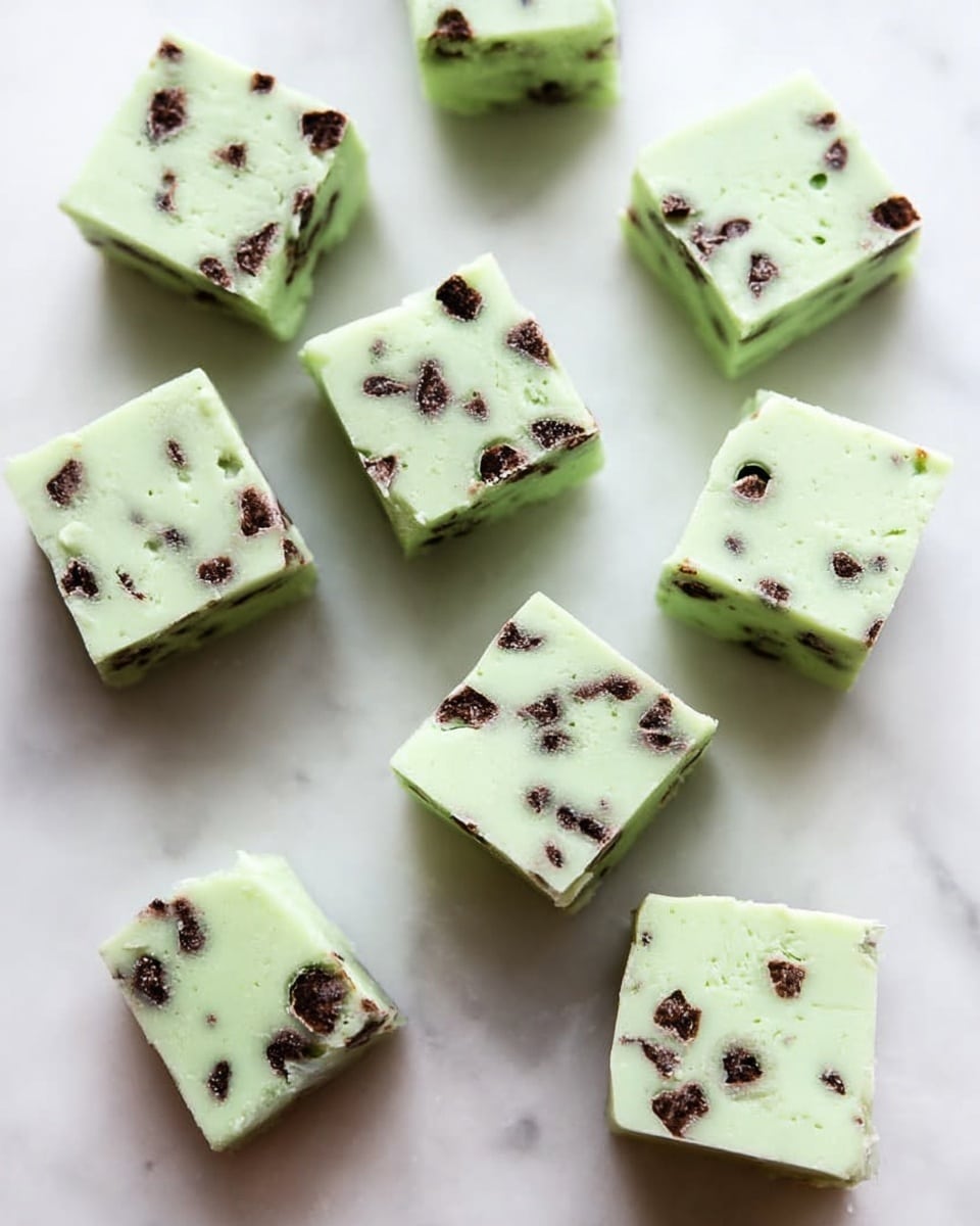 The image shows eleven small square pieces of light green fudge with tiny dark chocolate chips mixed evenly throughout each piece. The fudge pieces have smooth, slightly shiny surfaces and soft edges. They are spread out randomly on a white marbled surface, with no plate or bowl visible. The light green color and dark chips create a nice contrast, and the fudge looks thick and creamy. photo taken with an iphone --ar 4:5 --v 7