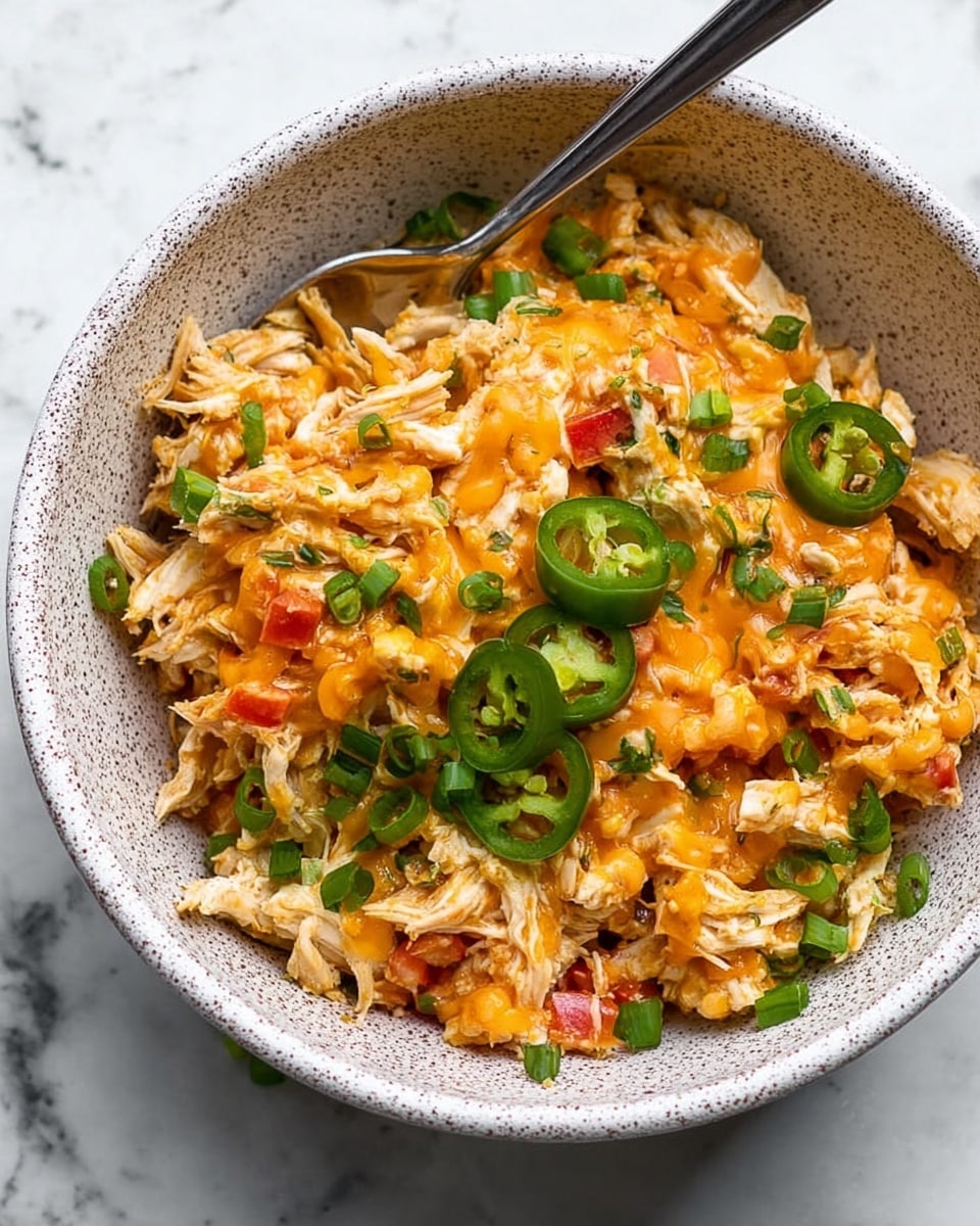 The dish is in a white speckled bowl filled with shredded cooked chicken as the main layer, mixed with small pieces of red bell pepper and diced onions, topped with bright orange melted cheese drizzled over the top. There are small green pea-like vegetables scattered throughout, and garnished with thinly sliced green jalapeño and chopped green onions. A silver spoon is placed inside the bowl, resting on one side, and the background is a white marbled texture. Photo taken with an iphone --ar 4:5 --v 7