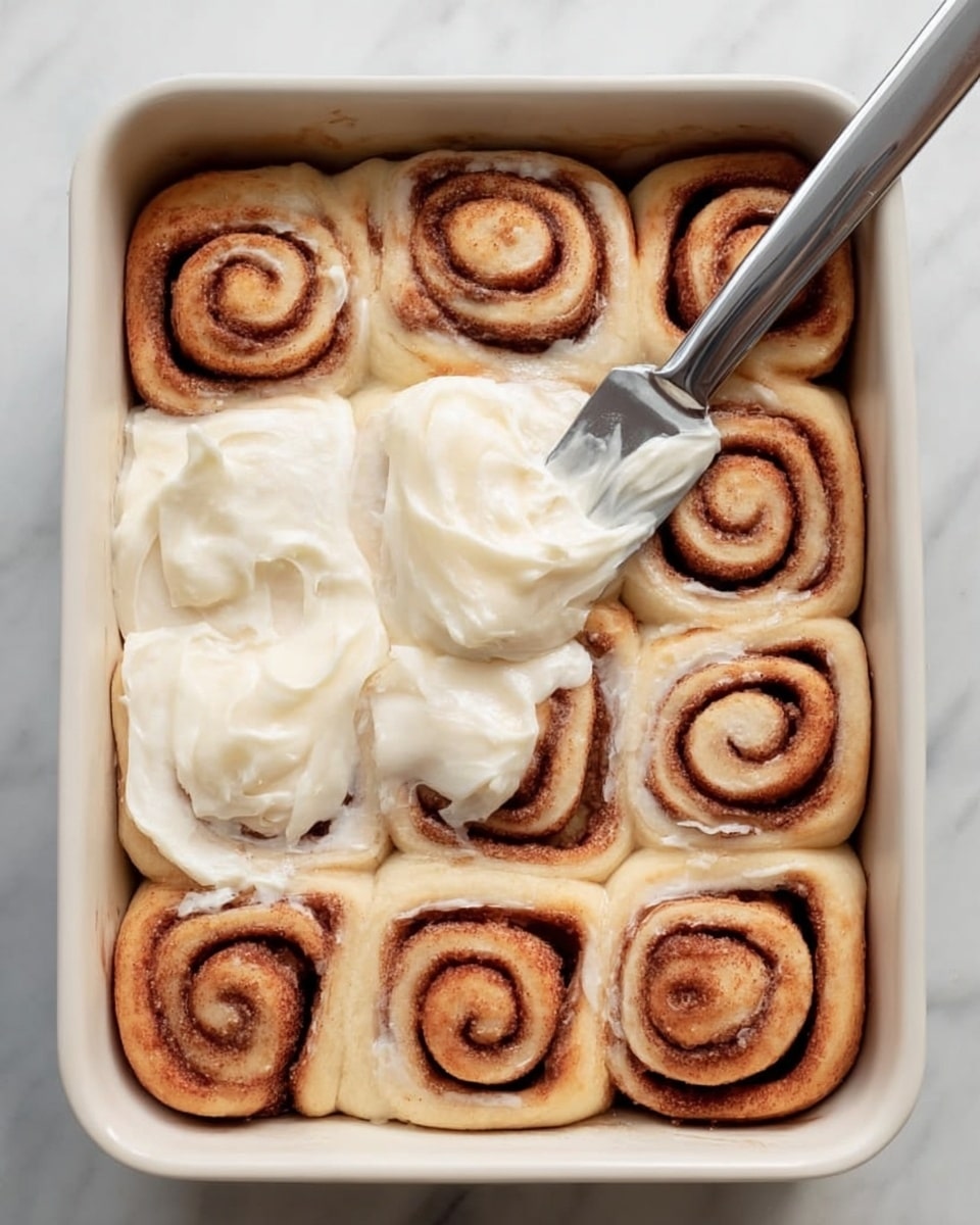 A white rectangular baking dish holds twelve cinnamon rolls arranged in three rows of four. Each roll has a light golden-brown dough with darker brown spiral cinnamon swirls. A woman's hand is spreading a thick layer of creamy white icing on the rolls with a silver spatula, focusing on the middle row. The dish is placed on a white marbled surface, with a soft blue cloth partially visible on the left side. Photo taken with an iphone --ar 4:5 --v 7