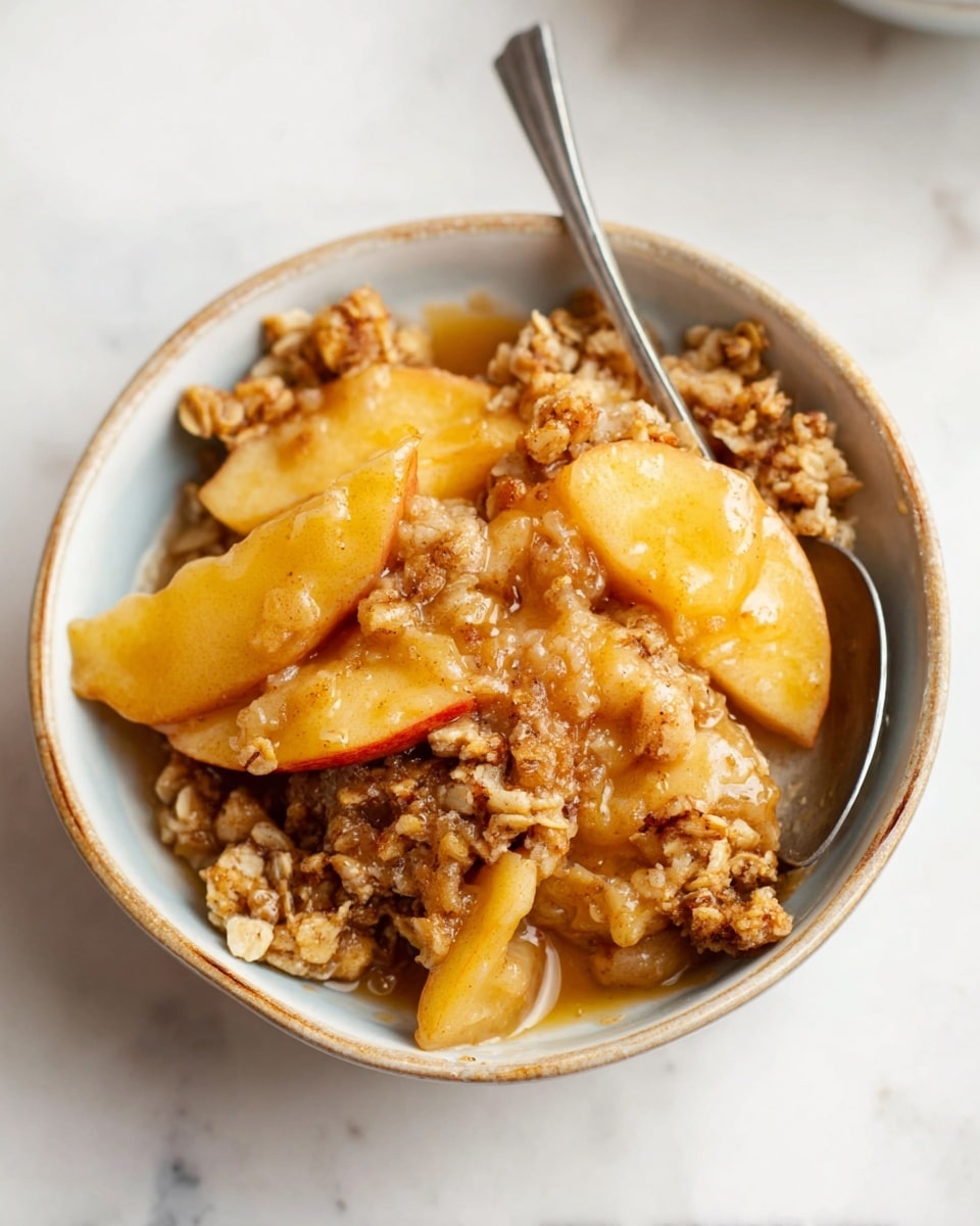 A small bowl filled with a warm apple crisp dessert showing two layers: the bottom layer with soft, light golden apple slices coated in a syrupy glaze, and the top layer with crumbly oat and brown sugar topping in uneven clusters, giving a crunchy texture. A silver spoon is partly inserted beside the apple slices, and the bowl sits on a white marbled surface. Photo taken with an iphone --ar 4:5 --v 7