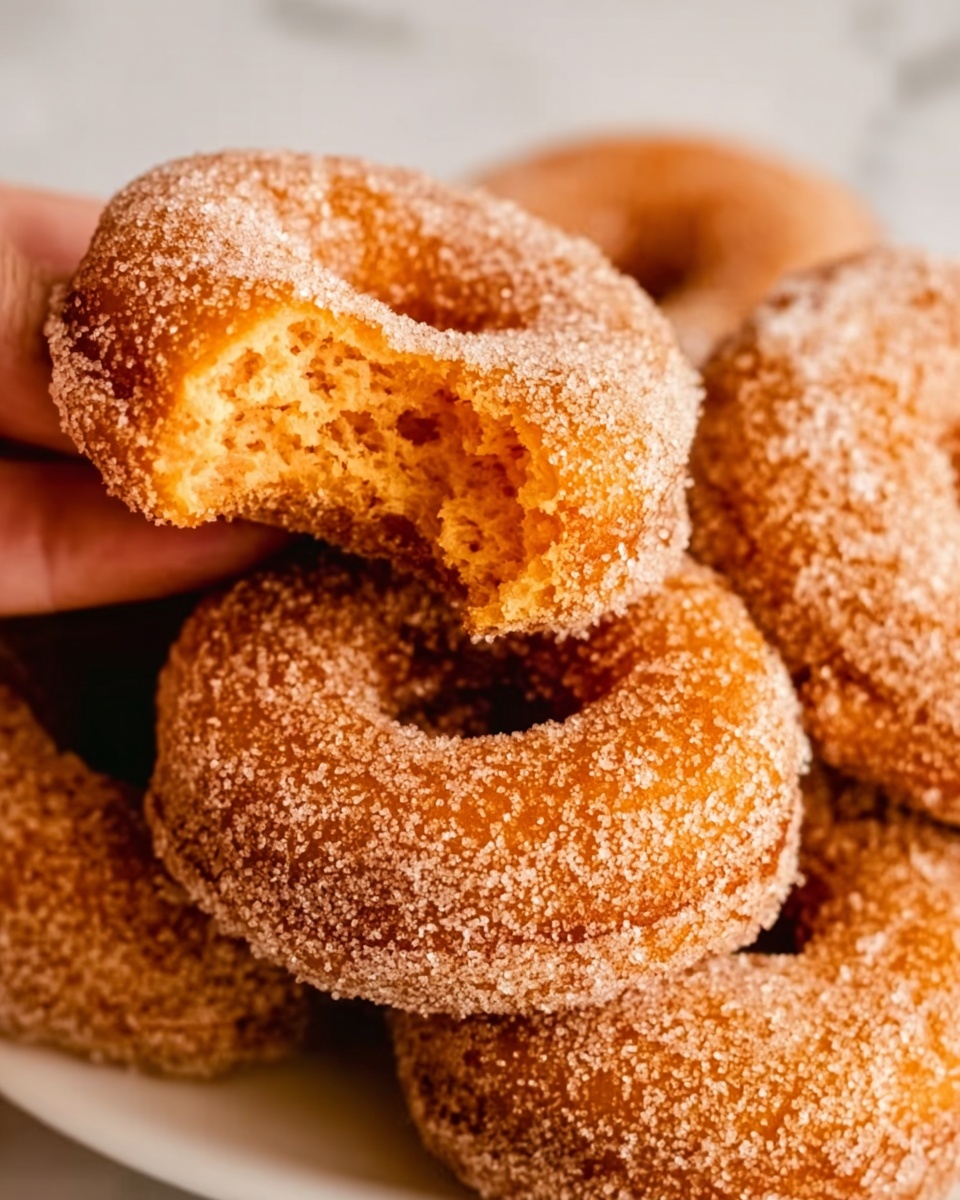 The image shows a close-up view of sugar-coated doughnuts stacked on top of each other. The top doughnut has a bite taken out, revealing a soft, orange-colored inside with a slightly rough texture. The doughnuts have a light brown, slightly crispy outside layer covered evenly with granulated sugar. The background is a white marbled surface. photo taken with an iphone --ar 4:5 --v 7