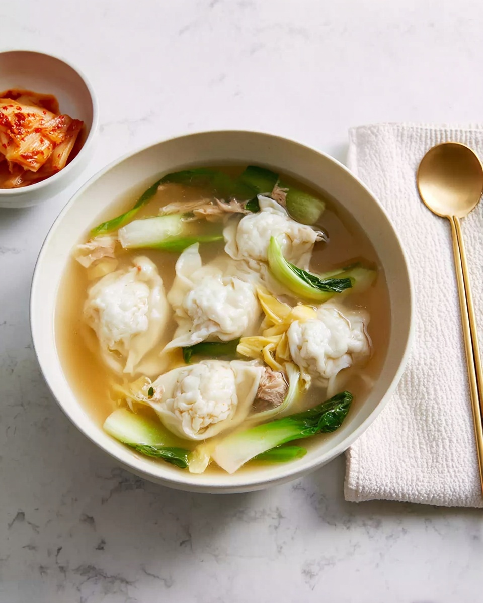 A white bowl filled with clear soup containing dumplings that are white and soft with slight folds, pieces of light beige cooked egg scattered around, bright green onion stalks partially submerged, and chunks of tender meat, all floating in a light brown broth. To the left is a smaller white bowl with reddish-orange kimchi showing a mix of cabbage and sauce. On the right, a white napkin with a gold spoon and gold chopsticks rests on a white marbled surface. Photo taken with an iphone --ar 4:5 --v 7