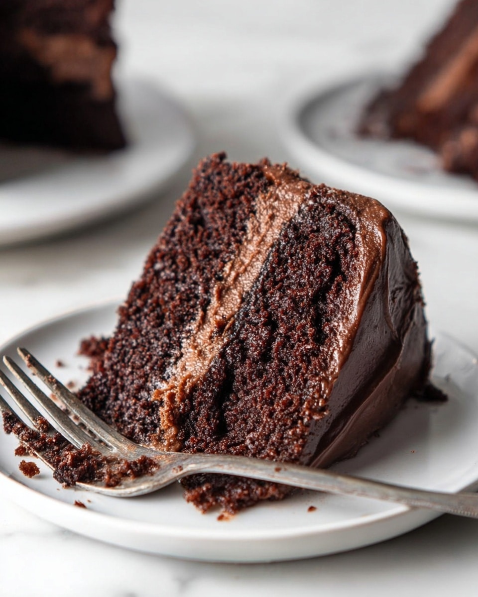 A close-up view of a slice of chocolate cake on a white plate showing two thick dark brown, moist layers with a creamy light brown chocolate filling layer in the middle and a smooth dark brown chocolate frosting coating the outside. The cake slice is slightly tilted, and there are small crumbs and chocolate bits scattered on the plate along with a metal fork placed in front of the slice. The scene is set on a white marbled surface with blurred out portions of two additional white plates with cake slices in the background. Photo taken with an iphone --ar 4:5 --v 7