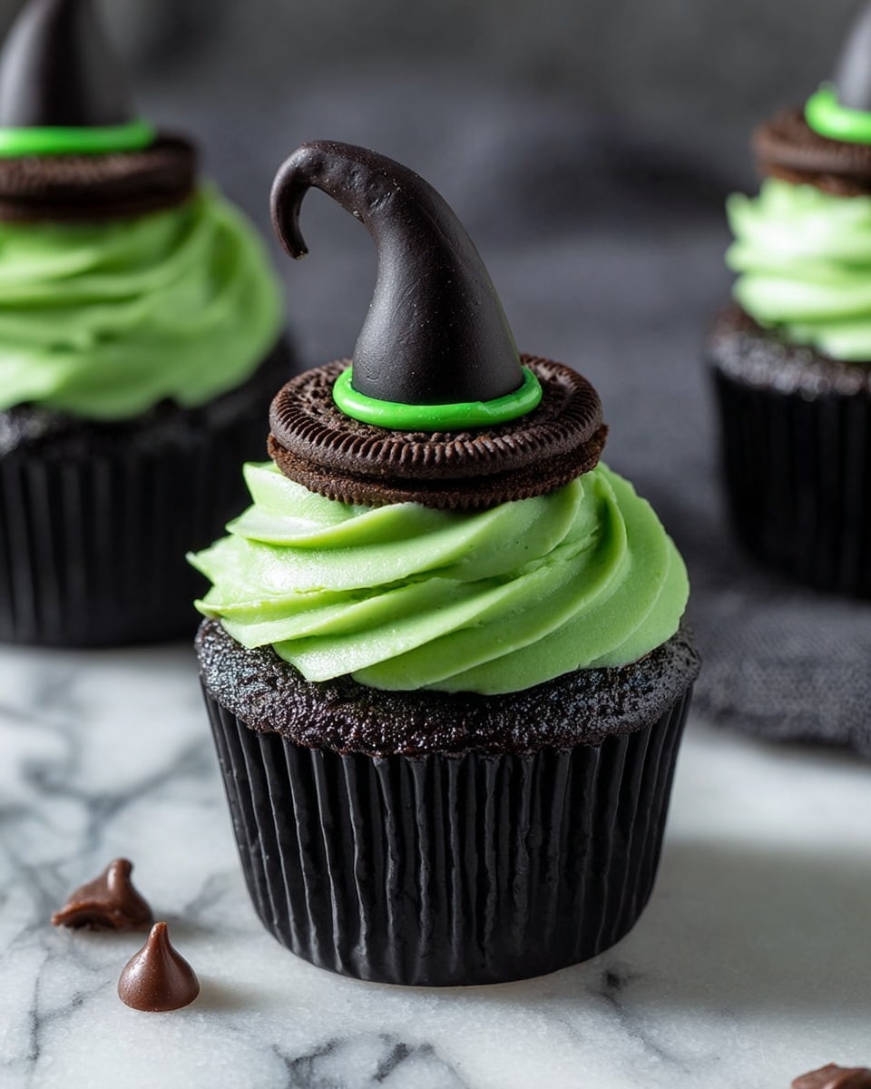 The image shows a dark chocolate cupcake with three layers: the base is a black cupcake wrapped in a black paper liner, the middle layer is a smooth, bright green swirled frosting, and the top layer is a chocolate cookie ring topped with a small, pointed black witch hat made of fondant with a green band at its base. The cupcake is placed on a white marbled surface with a few scattered chocolate chips around it. Photo taken with an iphone --ar 4:5 --v 7