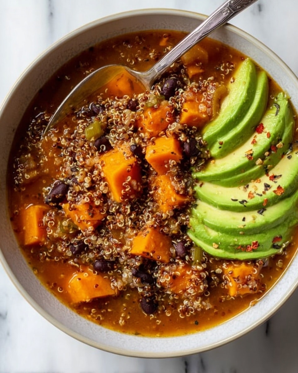 A close-up view of a bowl filled with a hearty mix of cooked quinoa, black beans, and bright orange chunks of sweet potato in a rich, slightly thick reddish broth, filling most of the bowl's inside. On one side of the bowl, there is a neatly sliced avocado with thin green layers slightly overlapping each other, sprinkled with black pepper flakes. A silver spoon rests inside the bowl, with its handle pointing outward, partially submerged in the quinoa and bean mixture. The bowl is white and sits on a white marbled surface. Photo taken with an iphone --ar 4:5 --v 7