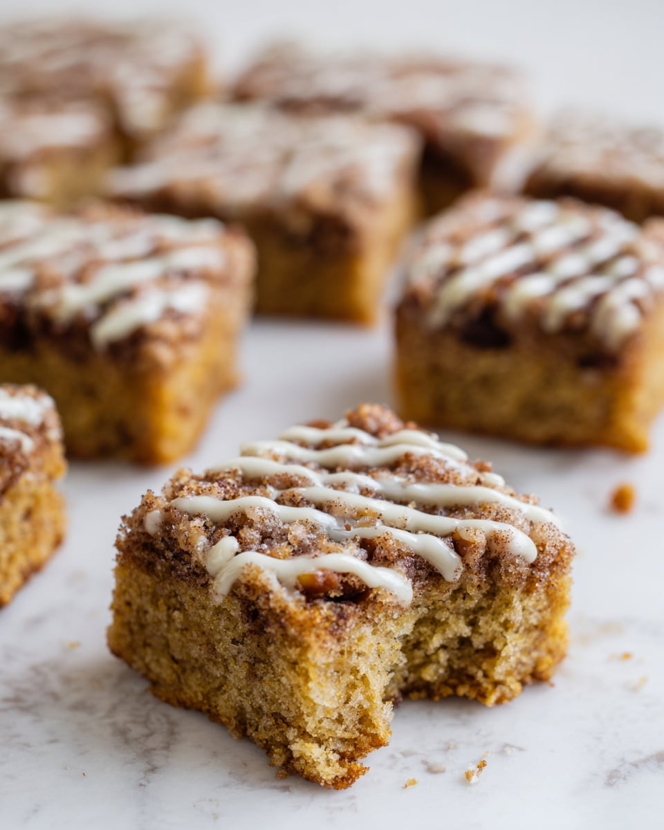 A close-up of several square pieces of a light brown cake arranged on a white marbled surface. Each piece is soft and moist looking with small dark brown spots throughout the cake body from nuts or chocolate. The top layer is a darker brown crumbly mixture with a white drizzle that adds texture and contrast. The piece in front has a bite taken out of it, showing the fluffy inside filled with small bits matching the top texture. Photo taken with an iphone --ar 4:5 --v 7