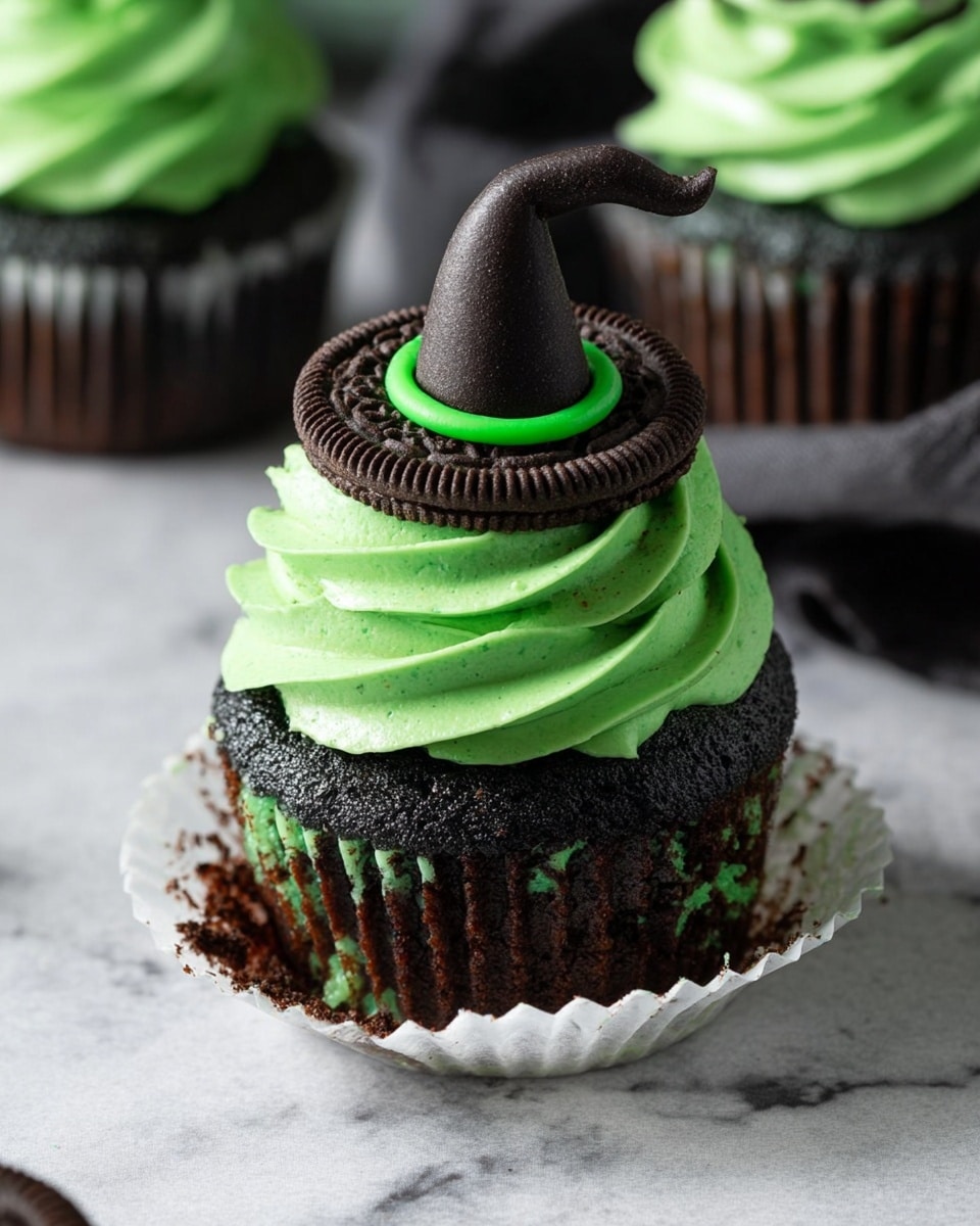 This image shows a dark black cupcake with chocolate chips inside, sitting in a peeled back white cupcake liner on a white marbled surface. The cupcake has a thick swirl of bright green frosting on top. On top of the frosting is a chocolate sandwich cookie with green frosting around its edge. A small black witch's hat made of fondant or similar material sits on top of the cookie with a green ring at its base. Another cupcake with the same decoration is blurred in the background. Photo taken with an iphone --ar 4:5 --v 7