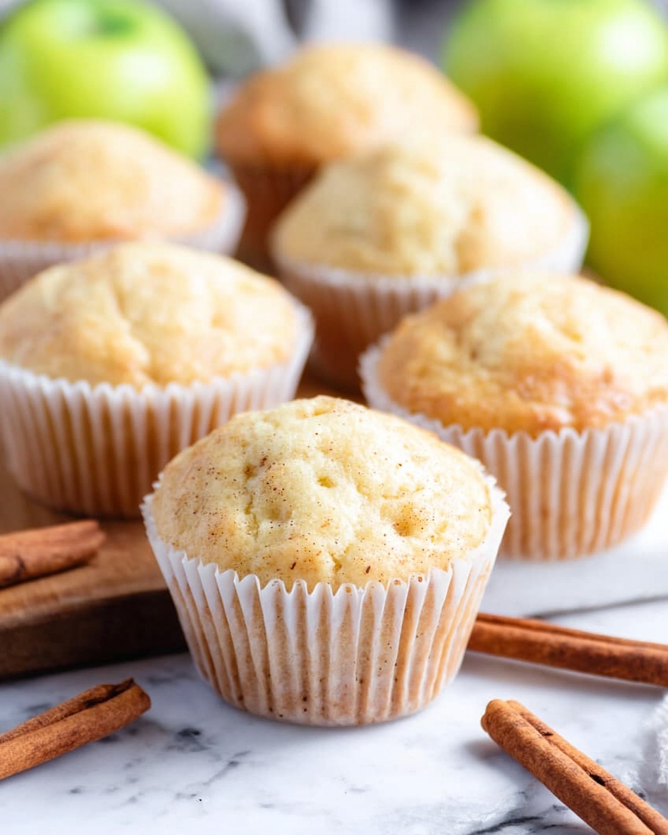 The image shows twelve light golden muffins with a slightly bumpy, soft texture, each wrapped in white paper liners. They are arranged mostly on an oval wooden board with a smooth, natural wood grain, sitting on a white marbled surface. Around the muffins, there are several whole green apples with smooth skin and a few cinnamon sticks scattered on the marble surface, adding a brown contrast. The scene feels fresh and cozy with natural lighting that highlights the muffins’ fluffy tops and the shiny apples. Photo taken with an iphone --ar 4:5 --v 7