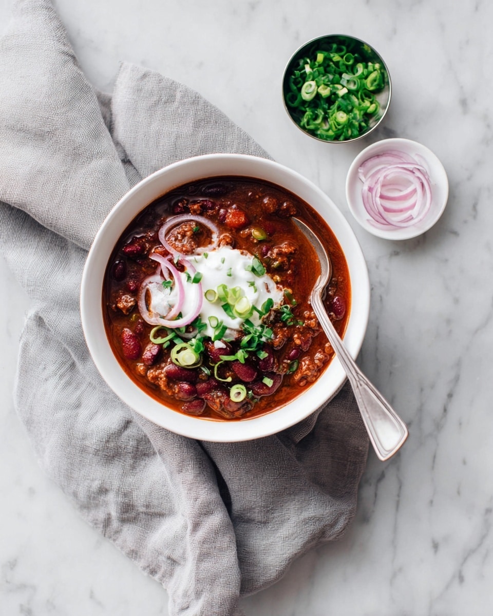 A white bowl holds a rich chili with visible kidney beans, ground meat, and tomato pieces in a deep red sauce. On top, there is a dollop of white sour cream, thin pale pink onion rings, and chopped green onions scattered over it. A silver spoon rests inside the bowl. The bowl is placed on a soft gray cloth, all set on a white marbled surface. Nearby, a small white dish has more thin onion rings, and a small reflective container holds extra chopped green onions. Photo taken with an iphone --ar 4:5 --v 7