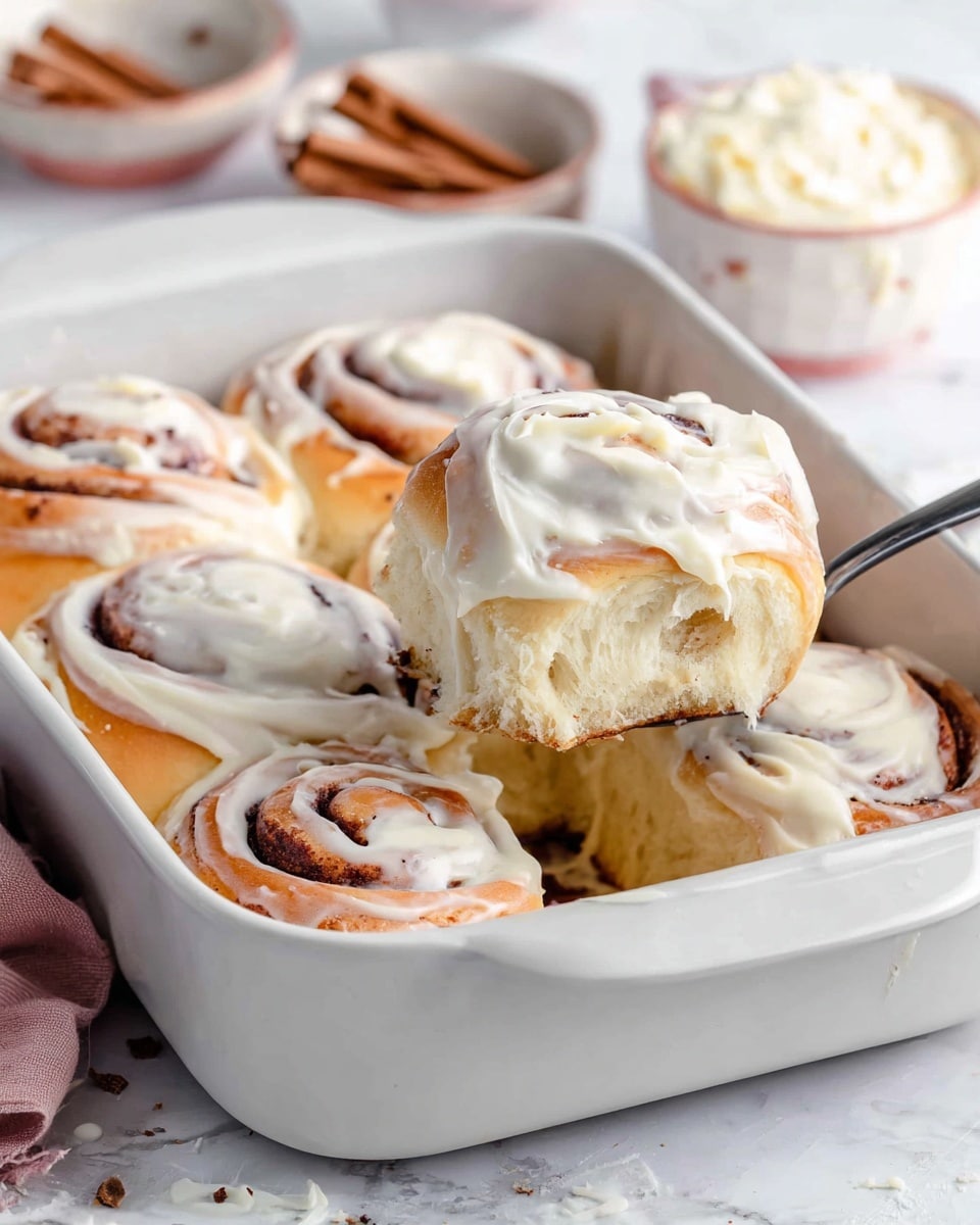 A white rectangular baking dish holds six soft, thick cinnamon rolls arranged in two rows, each roll showing a tight spiral of golden-brown dough with visible cinnamon layers. The top of each roll is covered in a creamy, white frosting that is thick but smoothly spread, dripping slightly over the edges. A silver utensil, lifted by a woman's hand, carries one of the rolls above the dish, revealing its fluffy, light interior with a slight sheen from the frosting. The dish rests on a white marbled surface, and soft background details include small bowls with cinnamon and frosting. Photo taken with an iphone --ar 4:5 --v 7