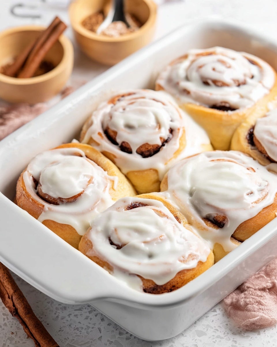 A single cinnamon roll sits in the middle of a white plate on a white marbled surface. The cinnamon roll has three visible layers of soft, light golden brown dough spiraled with a darker cinnamon filling inside. The top layer is thickly covered with creamy white icing that softly drapes over the sides and slightly pools around the base. In the background, there is another partially visible white plate with another cinnamon roll, slightly out of focus. A white baking dish with more cinnamon rolls is also partially visible nearby. Photo taken with an iphone --ar 4:5 --v 7