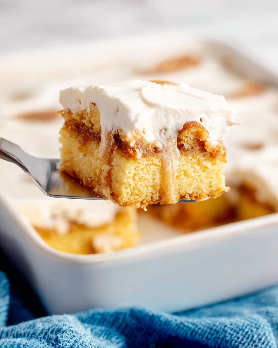 A close-up view of a square piece of cinnamon coffee cake being lifted by a metal spatula, showing two layers: the bottom layer is a soft yellow cake with a crumbly texture mixed with swirls of brown cinnamon filling, and the top layer is a smooth, thick white icing covering the cake evenly. The cake sits in a white baking dish on a white marbled surface with a folded blue cloth beside it. The background is softly blurred, keeping focus on the cake slice. Photo taken with an iphone --ar 4:5 --v 7