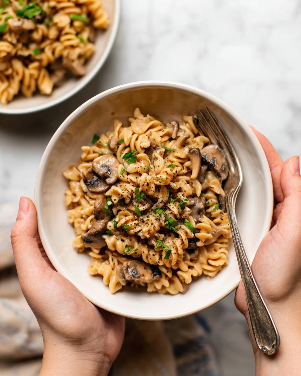 A close-up top view of creamy pasta with spiral-shaped noodles mixed with sliced brown mushrooms, all coated in a thick light brown sauce with visible specks of black pepper. The dish is inside a white pot, and there is a black and wooden spoon stirring the pasta on the right side. The surface around the pot is a white marbled texture. Photo taken with an iphone --ar 4:5 --v 7