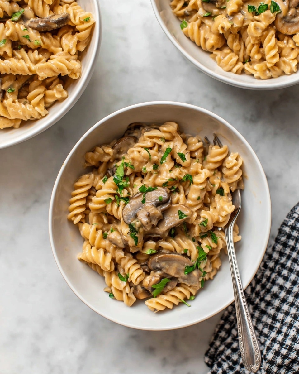 A white bowl filled with three layers visible: the bottom layer is creamy beige sauce coating light brown rotini pasta; the middle layer consists of cooked sliced mushrooms in medium brown shades mixed through the pasta; the top layer shows light green parsley leaves sprinkled and a few cracks of black pepper on the creamy pasta. A silver fork rests inside the bowl on the right side, partially inserted into the pasta. The bowl sits on a white marbled surface with part of a white and black checkered cloth visible at the bottom right. Photo taken with an iphone --ar 4:5 --v 7