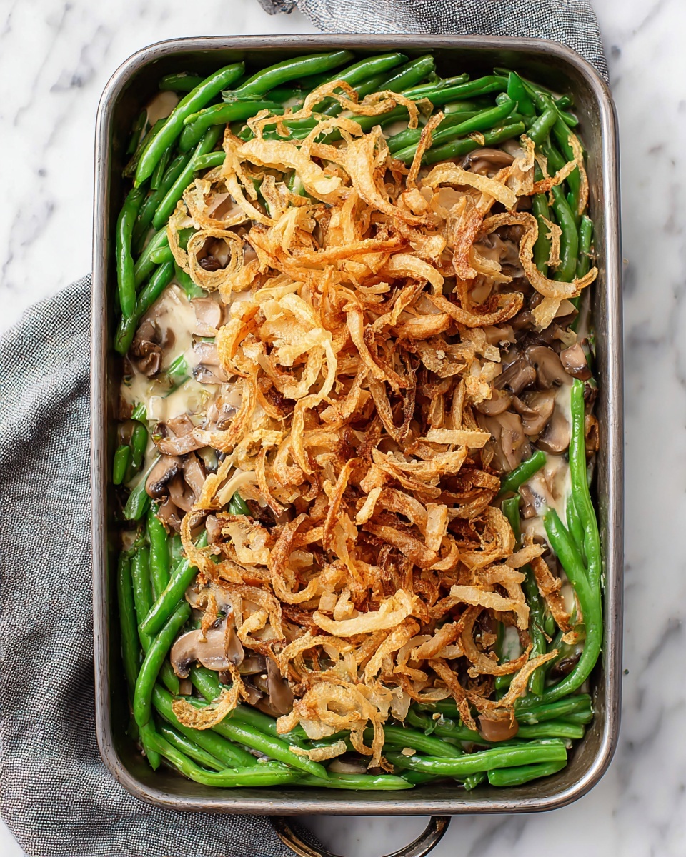A rectangular metal baking dish filled with a green bean casserole, showing three main layers. The bottom layer is whole green beans, bright green and lined up to cover the base. On top of the green beans is a light brown mushroom sauce with visible mushroom slices scattered unevenly. The top layer consists of crispy, thin fried onion strips, golden brown with some darker edges, piled high in the center and spilling slightly over the sides. The dish sits on a white marbled surface with a gray cloth nearby. Photo taken with an iphone --ar 4:5 --v 7
