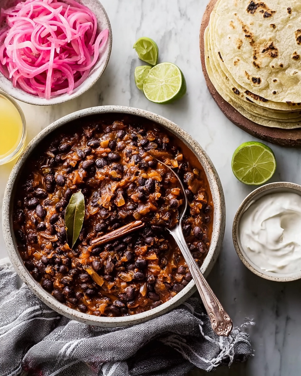 The image shows a bowl filled with black bean stew that has a thick, chunky texture with visible beans and diced vegetables mixed with a reddish-brown sauce, garnished with bay leaves and a cinnamon stick on top. A spoon is resting inside the bowl, which is placed on a white marbled surface next to a white bowl containing bright pink pickled onions. On the right side, there is a small stack of round, lightly grilled white tortillas arranged loosely, with lime wedges scattered nearby. A small white bowl of smooth sour cream with a spoon is also present, along with two glasses of a light yellow drink. A beige cloth napkin with a dark stripe is tucked under the bowl of stew, and the whole scene is well-lit, natural, and cozy. Photo taken with an iphone --ar 4:5 --v 7