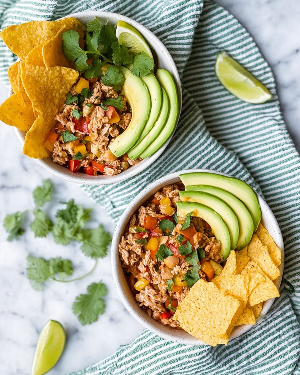 Two white bowls on a white marbled surface, each filled with three main sections: shredded light brown chicken mixed with small red and yellow bell pepper pieces and green cilantro leaves in the bottom left, golden yellow tortilla chips stacked on the right side, and thin slices of green avocado fanned out on the left edge. A small wedge of lime with a bright green peel rests on top of the chicken mix in each bowl. The bowls are placed near a green and white striped cloth, with a few lime wedges and cilantro leaves scattered around. photo taken with an iphone --ar 4:5 --v 7