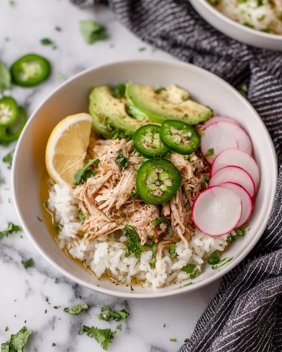 A white bowl holds a dish with three visible layers: the bottom layer is a bed of white rice with a slightly grainy texture, topped by shredded light brown chicken pieces that look tender and moist spread across the center, and the top layer includes thin slices of avocado in pale green, bright green jalapeno rings, and two round, thin slices of radish with white centers and pink edges on the right side. A small wedge of lemon with a yellowish color sits on the left side, and small green cilantro leaves are sprinkled over the top. The bowl is set on a white marbled surface next to a black and white striped cloth, with some scattered green herbs around. photo taken with an iphone --ar 4:5 --v 7