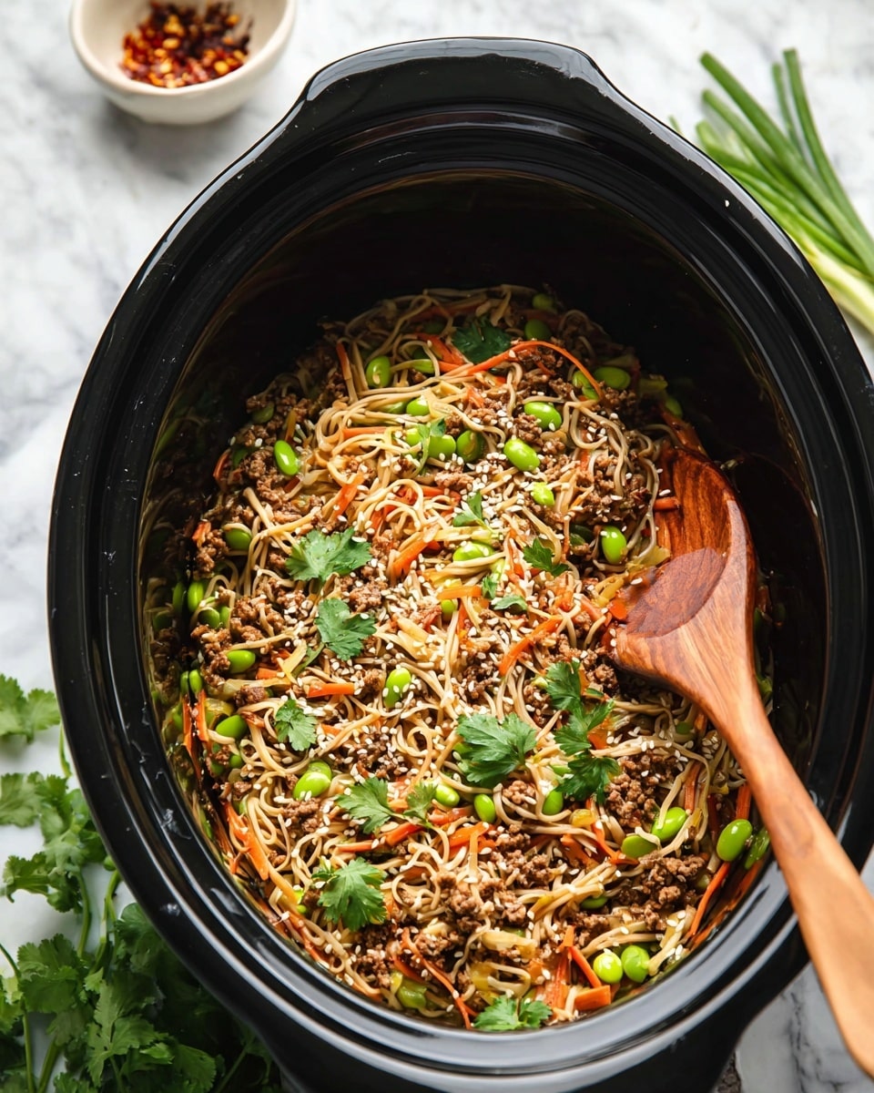 Inside a black slow cooker, there is a mixed noodle dish with thin, light beige noodles layered with small brown ground meat pieces, thin orange carrot strips, and bright green edamame beans and sliced green onions sprinkled throughout. The dish is garnished with fresh green cilantro leaves and white sesame seeds. A wooden spoon rests on the right side, partially buried in the noodles. The background is a white marbled surface with some green onion pieces and a small white bowl of red chili flakes visible around the cooker. Photo taken with an iphone --ar 4:5 --v 7