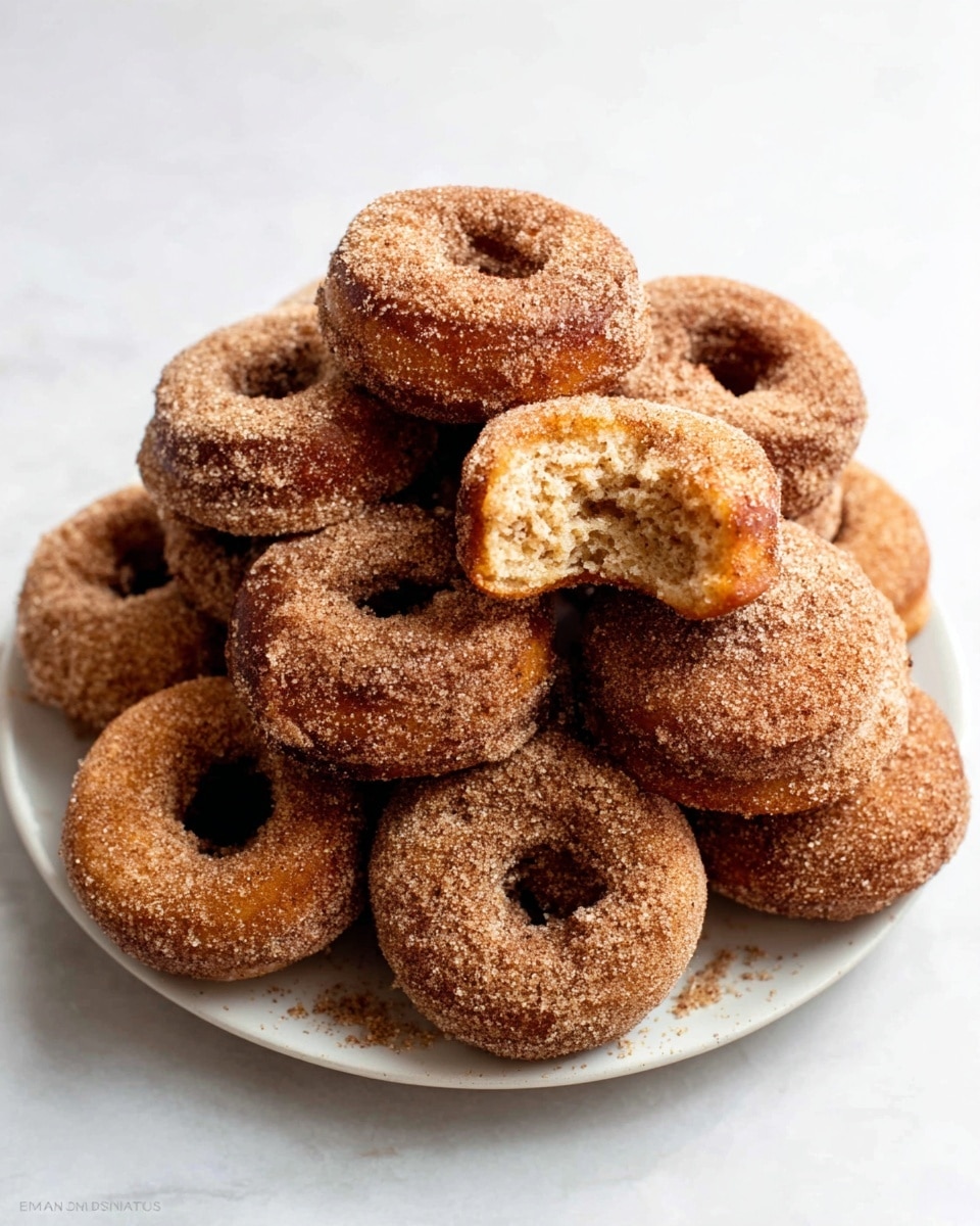 The image shows a stack of four round doughnuts covered in a fine layer of sugar with a brown, slightly rough texture. Three doughnuts are stacked on top of each other, and one is leaning against the stack on the right side. To the left, there are a few pieces of broken doughnut pieces that reveal a light golden interior with a soft and airy texture. The doughnuts sit on a white marbled surface with a clean, bright background. Photo taken with an iphone --ar 4:5 --v 7