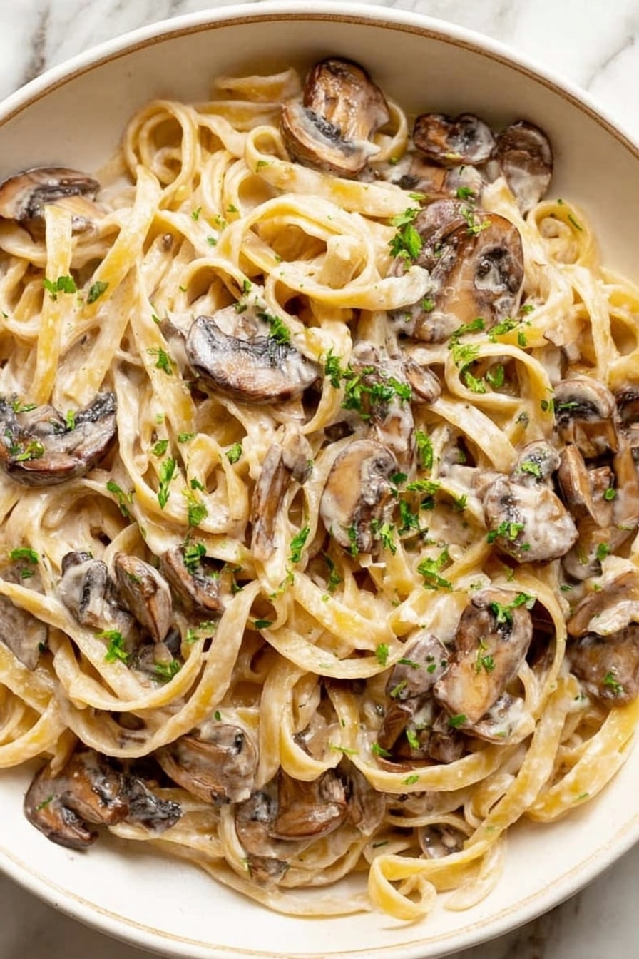 This image shows a white plate filled with creamy pasta mixed with cooked mushroom slices. The pasta is light golden and twisted, with a smooth creamy sauce coating each strand. Dark brown mushrooms are spread throughout, some partially covered by the sauce. Small green herbs are sprinkled on top, adding a fresh color contrast against the creamy beige sauce and pasta. The plate sits on a white marbled surface, and the overall look is warm and inviting. photo taken with an iphone --ar 4:5 --v 7