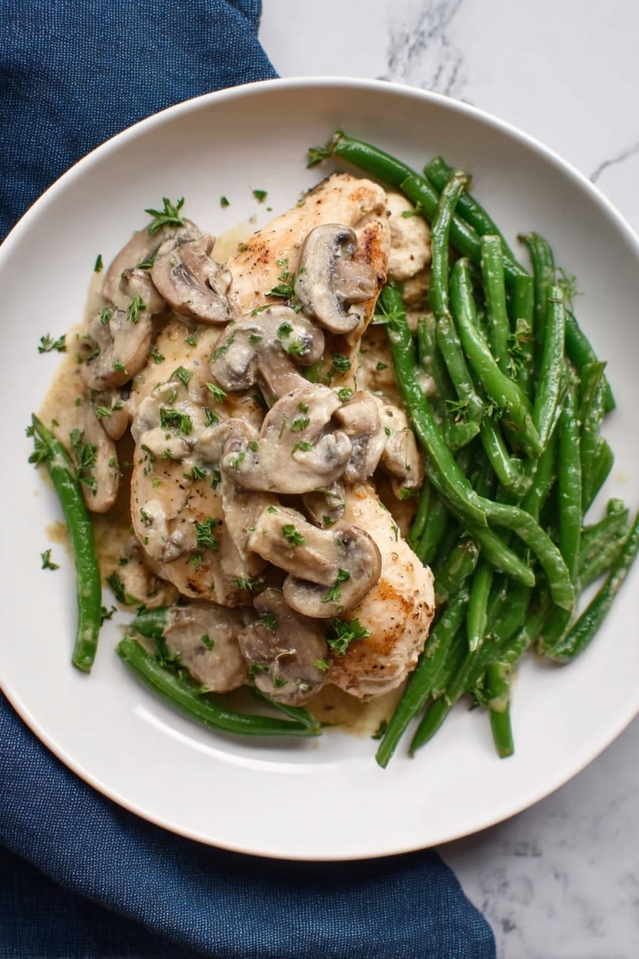 Easy Chicken Marsala Without Wine Recipe 6 The dish shows two pieces of light brown cooked chicken placed on a layer of green beans with a slightly shiny texture, all on a white plate. On top of the chicken and green beans are many light tan sliced mushrooms in a creamy sauce that looks soft and smooth. Fresh green chopped herbs are sprinkled over the chicken, mushrooms, and beans, adding a bright contrast. The plate sits on a white marbled surface with a dark blue cloth nearby. photo taken with an iphone --ar 4:5 --v 7