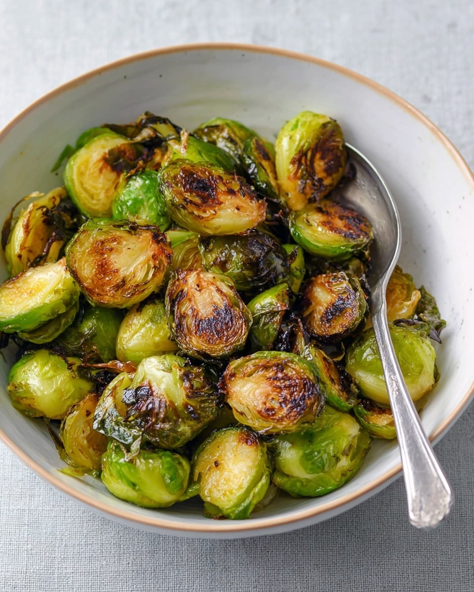 A white bowl is filled with cooked Brussels sprouts that have a mix of bright green and dark brown crispy leaves. Some sprouts are sliced in half showing a soft-looking pale green inside with a golden brown surface, while others are whole or loose leaves with a crunchy texture. The bowl sits on a white marbled surface with a silver spoon partially inside the bowl on the right side. The overall look shows a mix of soft and crunchy textures with warm browned spots on the green vegetables photo taken with an iphone --ar 4:5 --v 7