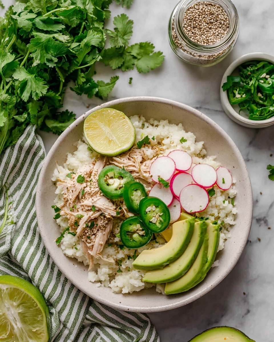 A white bowl filled with a base layer of white cauliflower rice, topped with shredded light brown chicken in the center. On top of the chicken, there are thin green jalapeño slices scattered, along with three thin white and pink radish slices on one side and three light green avocado slices on the other. A small wedge of yellow lime is placed on top near the back edge of the bowl. Fresh green cilantro leaves are sprinkled all around. The bowl is set on a white marbled surface, next to a bundle of fresh cilantro, a green and white striped cloth, a green-lidded jar of seeds, and half a lime, photo taken with an iphone --ar 4:5 --v 7