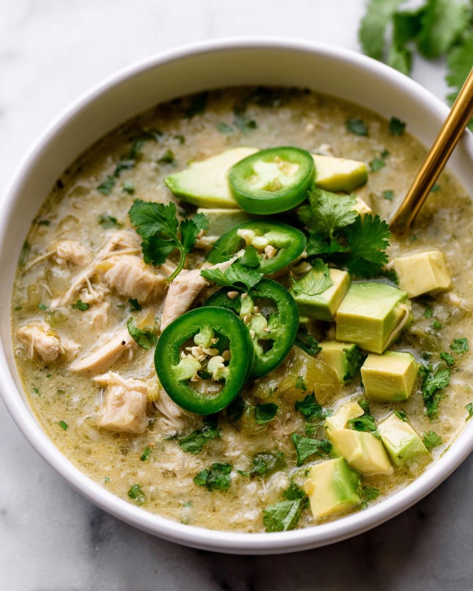 The image shows a white bowl filled with a thick, light green soup. Inside the soup, there are chunks of light beige cooked chicken and visible bits of chopped herbs. On top, there are thin slices of bright green jalapeño peppers placed in a row, along with small cubes and slices of fresh yellow-green avocado. Fresh green cilantro leaves are scattered on the right side of the bowl, adding a leafy texture. A gold spoon is partially immersed in the soup on the right side of the bowl. The bowl is set on a white marbled surface. Photo taken with an iphone --ar 4:5 --v 7