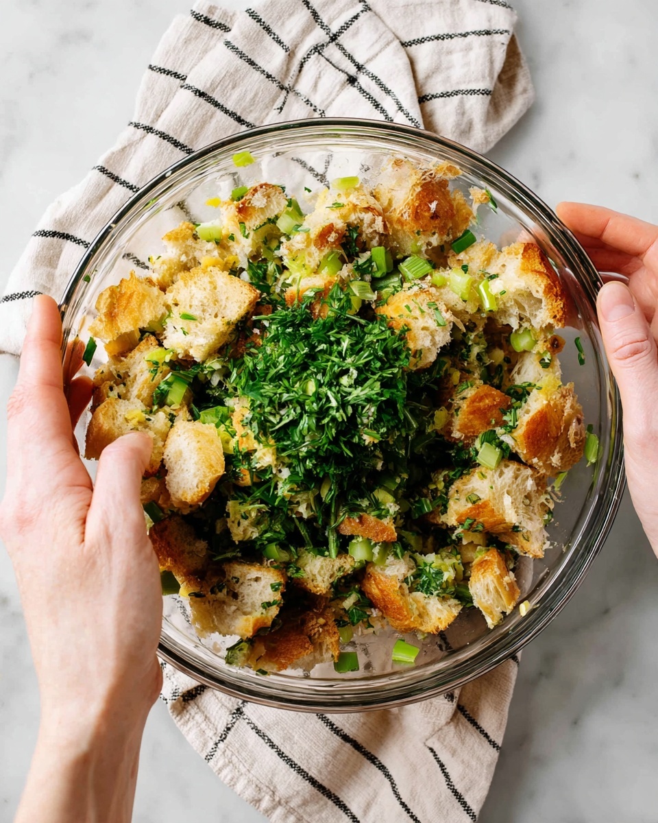 A clear glass bowl filled with roughly torn pieces of toasted bread, showing a mix of golden brown crusts and soft white insides. Scattered over the bread are chopped light green celery and bright green herbs, adding fresh color and texture throughout the bowl. A wooden spoon rests inside the bowl on the right side, slightly nestled among the bread and greens. The bowl sits on a white marbled surface with a lightly patterned cloth folded nearby. Photo taken with an iphone --ar 4:5 --v 7