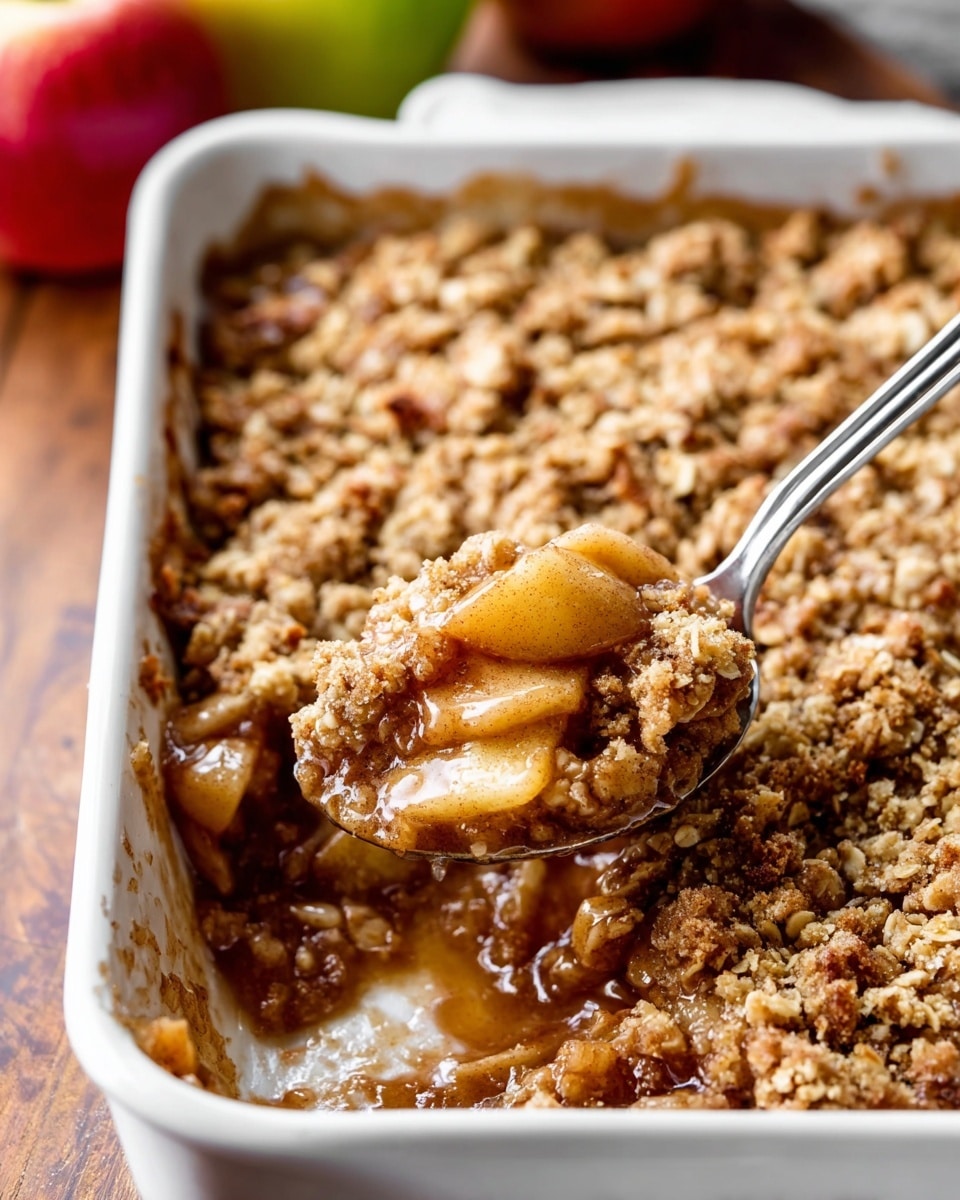 This image shows a white baking dish filled with apple crumble. The bottom layer is soft, brownish apple chunks with a glossy, syrupy texture. The top layer is thick and crumbly with a mix of oats and golden brown streusel. A shiny silver spoon is scooping out some of the apple filling with crumble, positioned near the right side of the dish. The dish sits on a wooden surface, and there are blurred apples nearby. The photo taken with an iphone --ar 4:5 --v 7