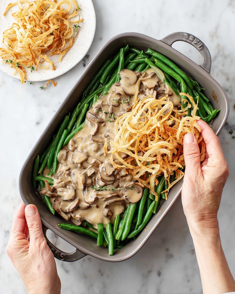 The image shows a casserole dish with three clear layers. The bottom layer has fresh green beans arranged neatly around the edges, with bright green color and smooth texture. The middle layer is a creamy mushroom sauce, light beige in color, covering the center in a thick spread with visible mushroom slices. On top, there is a pile of golden-brown crispy fried onions being placed by a woman's hands, with the onions having a thin, curly texture and some darker browned edges. To the side, there is a small white wooden plate with extra fried onions. The background is a white marbled surface. Photo taken with an iphone --ar 4:5 --v 7
