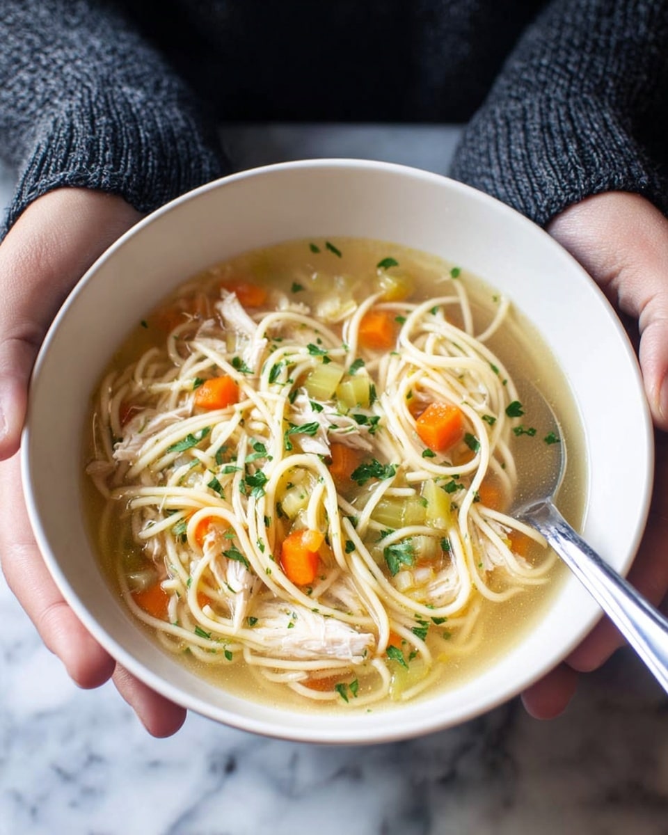 A bowl of clear chicken noodle soup is held by a pair of woman's hands wearing a dark gray long sleeve sweater. Inside the white bowl, there are several layers: a light broth base with thin yellow noodles floating, mixed with bright orange carrot slices, small pieces of light green celery, and shredded white chicken meat. Fresh green herbs are sprinkled on top, adding a touch of color contrast. A metal spoon rests on the right side inside the bowl, partially submerged in the broth. The photo is taken against a white marbled surface. photo taken with an iphone --ar 4:5 --v 7