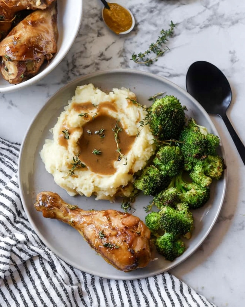A white plate holds two golden-brown cooked drumsticks placed side by side at the lower center. Above the drumsticks is a round scoop of creamy mashed potatoes with a crater filled with brown gravy, topped with green herb sprigs. On the right side of the plate, there is a cluster of bright green roasted broccoli florets. The plate sits on a white marbled surface next to a black spoon and a white cloth with black stripes. In the top left corner, there is a white bowl with more drumsticks and a small amount of mustard next to a spoon. photo taken with an iphone --ar 4:5 --v 7