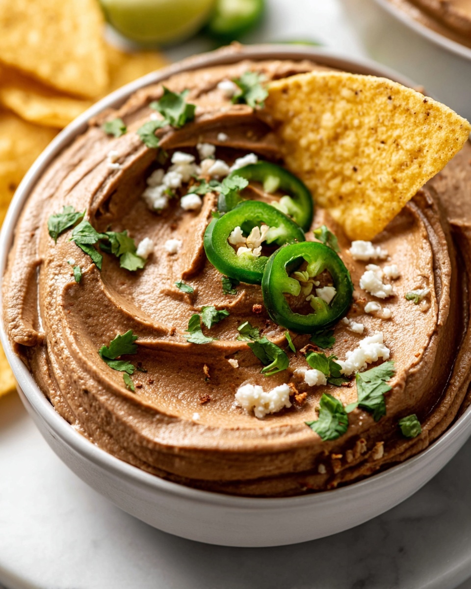 Crockpot Refried Beans Recipe 5 A white bowl filled with smooth brown dip, swirled across the surface in thick, creamy layers. On top, there are small white crumbly bits and scattered green cilantro leaves, with a thin green jalapeño slice adding sharp contrast. A yellow tortilla chip is partially dipped in the brown dip, resting on the edge of the bowl. The bowl sits on a white marbled surface with a corner of another white bowl visible in the background. Photo taken with an iphone --ar 4:5 --v 7