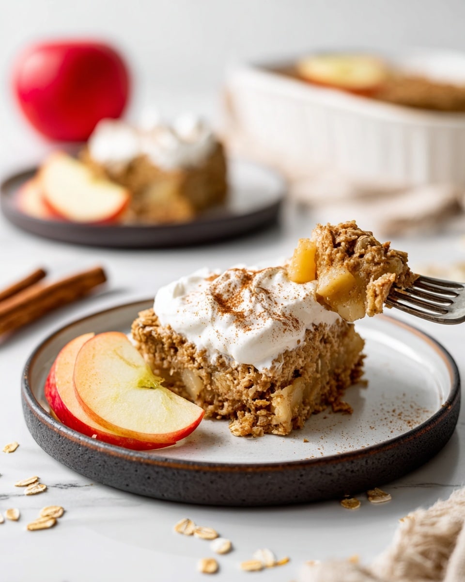 A piece of baked oatmeal sits on a round white plate with a dark rim, topped with a dollop of white cream sprinkled with light brown cinnamon. The oatmeal is golden brown with visible small chunks of fruit inside, showing a soft and crumbly texture. Next to the oatmeal, two apple slices with red and yellow skin rest on the plate. A silver fork holds a small bite of the oatmeal, with cream on top. In the background, a whole red apple with yellow streaks, a white bowl with more yellow fruit pieces, and another plate of oatmeal are placed on a white marbled surface. There are scattered oatmeal flakes and a peach-colored cloth nearby. Photo taken with an iphone --ar 4:5 --v 7