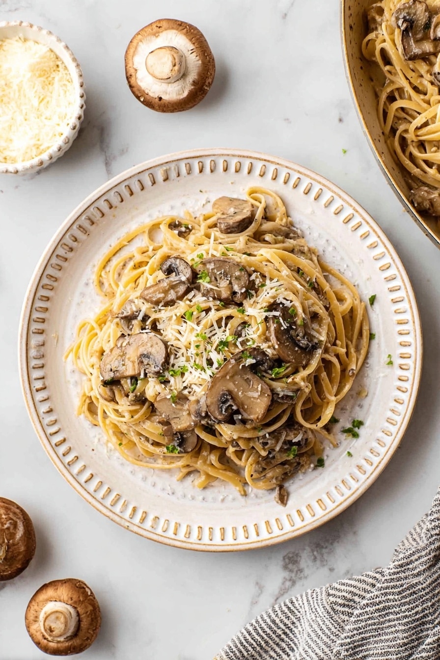 A white round plate with small round beads on the rim holds two layers of fettuccine pasta mixed with creamy mushroom sauce. The pasta is light brown with smooth texture, and the mushrooms are sliced, dark brown, and scattered evenly throughout. The dish is topped with small flakes of white cheese and sprinkled with chopped green herbs. Around the plate are whole and sliced mushrooms and a small white bowl with pale yellow cheese slices on a white marbled surface, next to a striped cloth napkin. Photo taken with an iphone --ar 4:5 --v 7