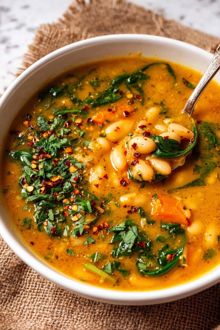 A white bowl filled with a thick orange-yellow soup containing white beans, small orange carrot pieces, and green spinach leaves floating throughout. The soup is topped with scattered red chili flakes and chopped fresh green herbs. A spoon inside the bowl holds a mix of beans and carrots in the soup. The bowl sits on a coarse brown burlap cloth on a white marbled surface. Photo taken with an iphone --ar 4:5 --v 7
