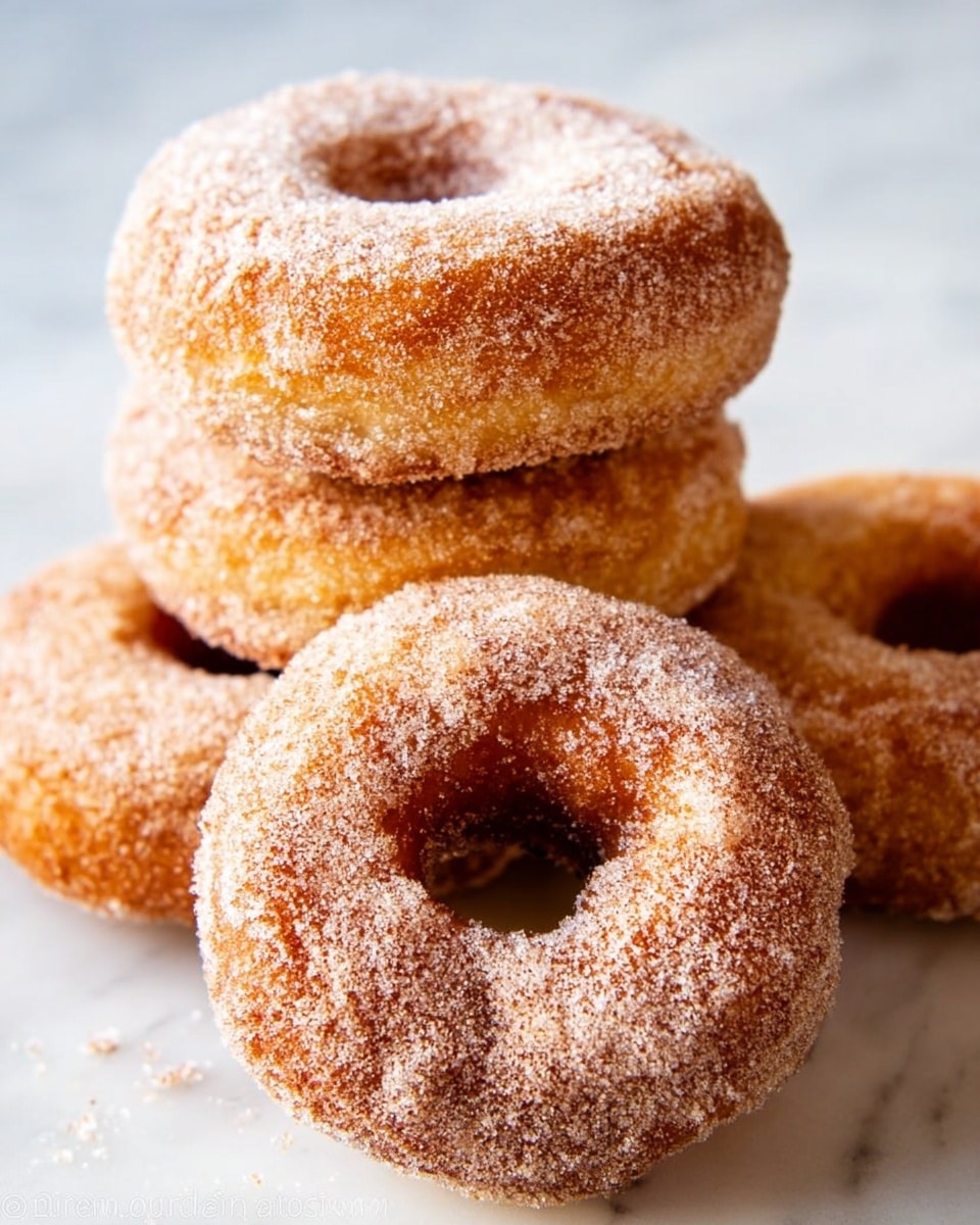 The image shows several round donuts on a white marbled surface, with a golden-brown color and a coating of granulated sugar mixed with cinnamon dusted all over. One donut is broken in half, revealing a soft, light, fluffy inside with a slightly crumbly texture. The donuts have a rough, slightly crispy outer layer with visible sugar crystals. The donuts are arranged close together, showing the warm tones of the sugar and dough clearly. Photo taken with an iphone --ar 4:5 --v 7
